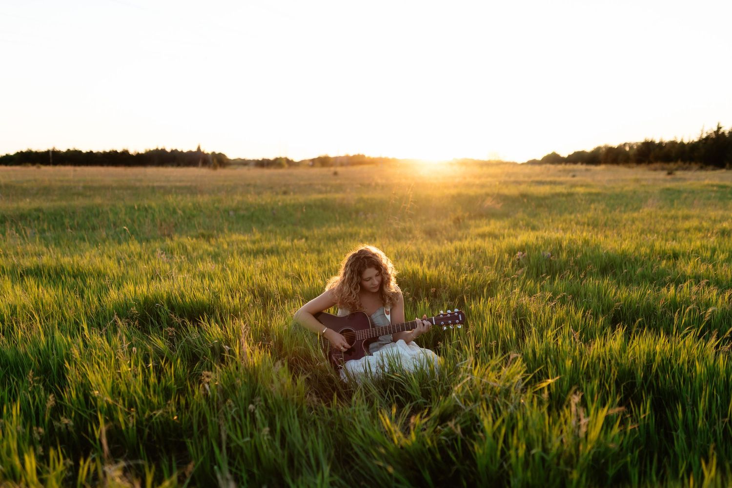 Woman playing acoustic guitar in a sunlit green field at golden hour sunset.