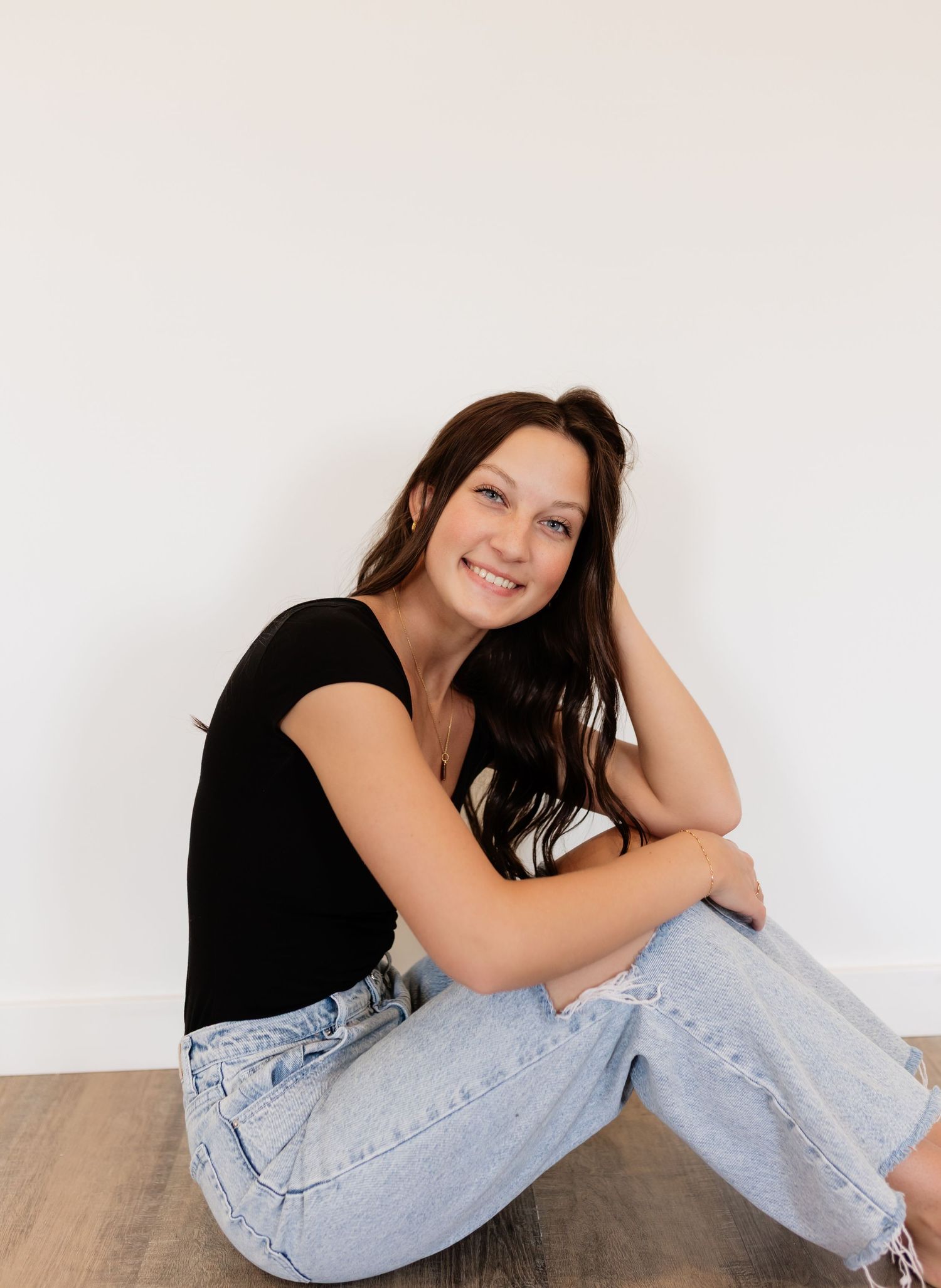 Young woman in black tee and light jeans sitting on floor smiling casually.