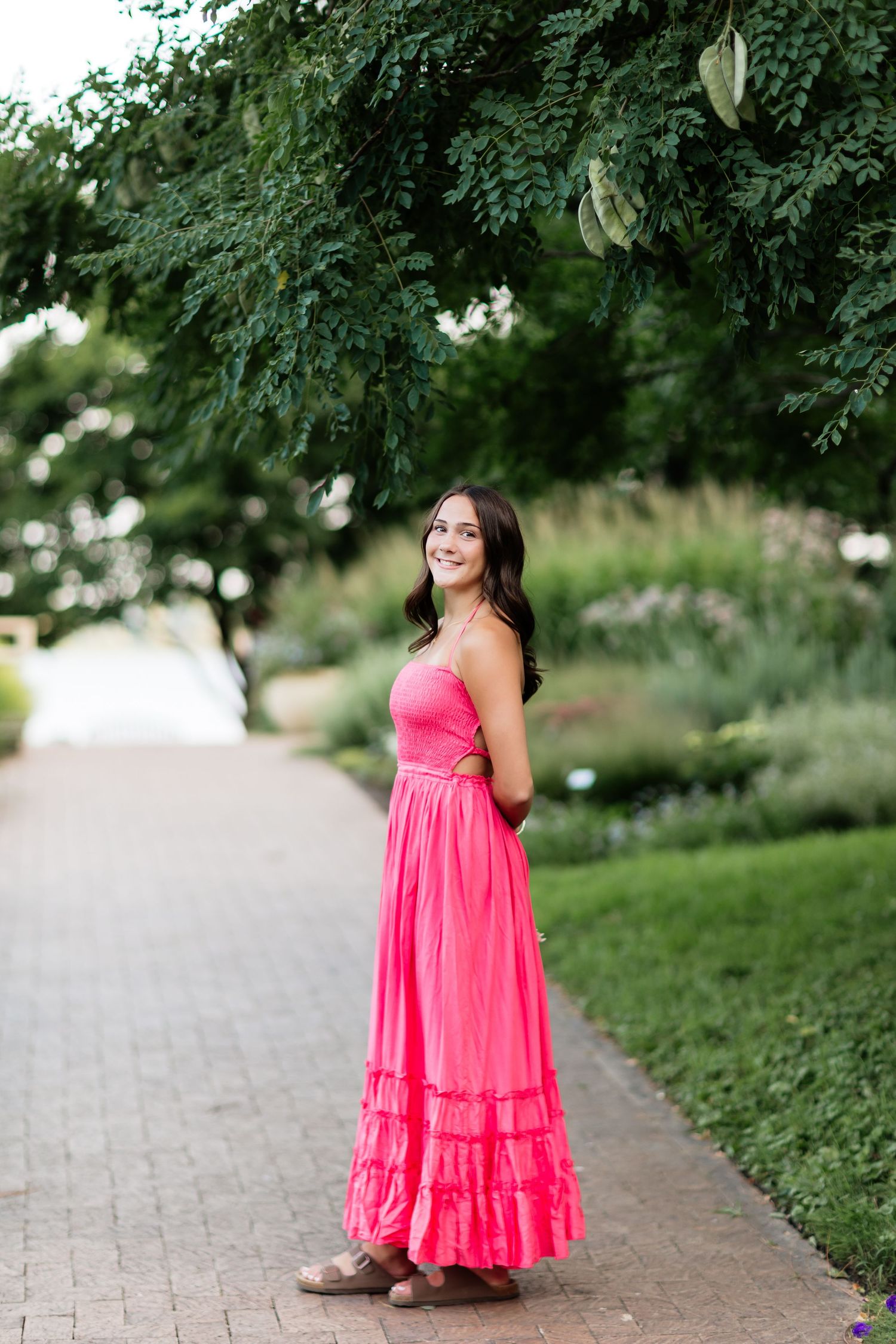 Young woman in a bright pink sleeveless maxi dress smiling while standing on a brick path in a lush garden.