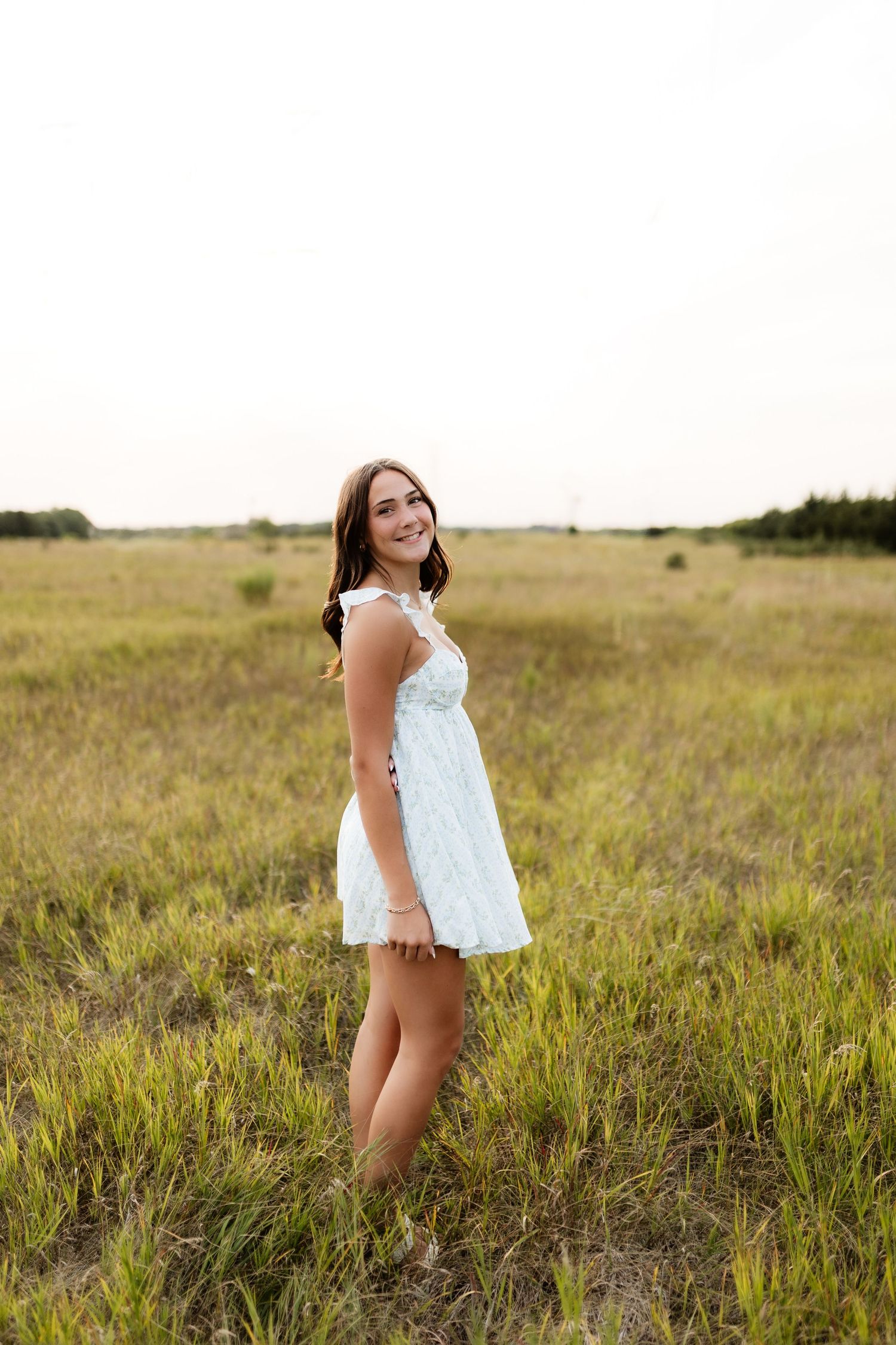 Young woman in white sundress smiling in a green field, posing outdoors on a bright day.