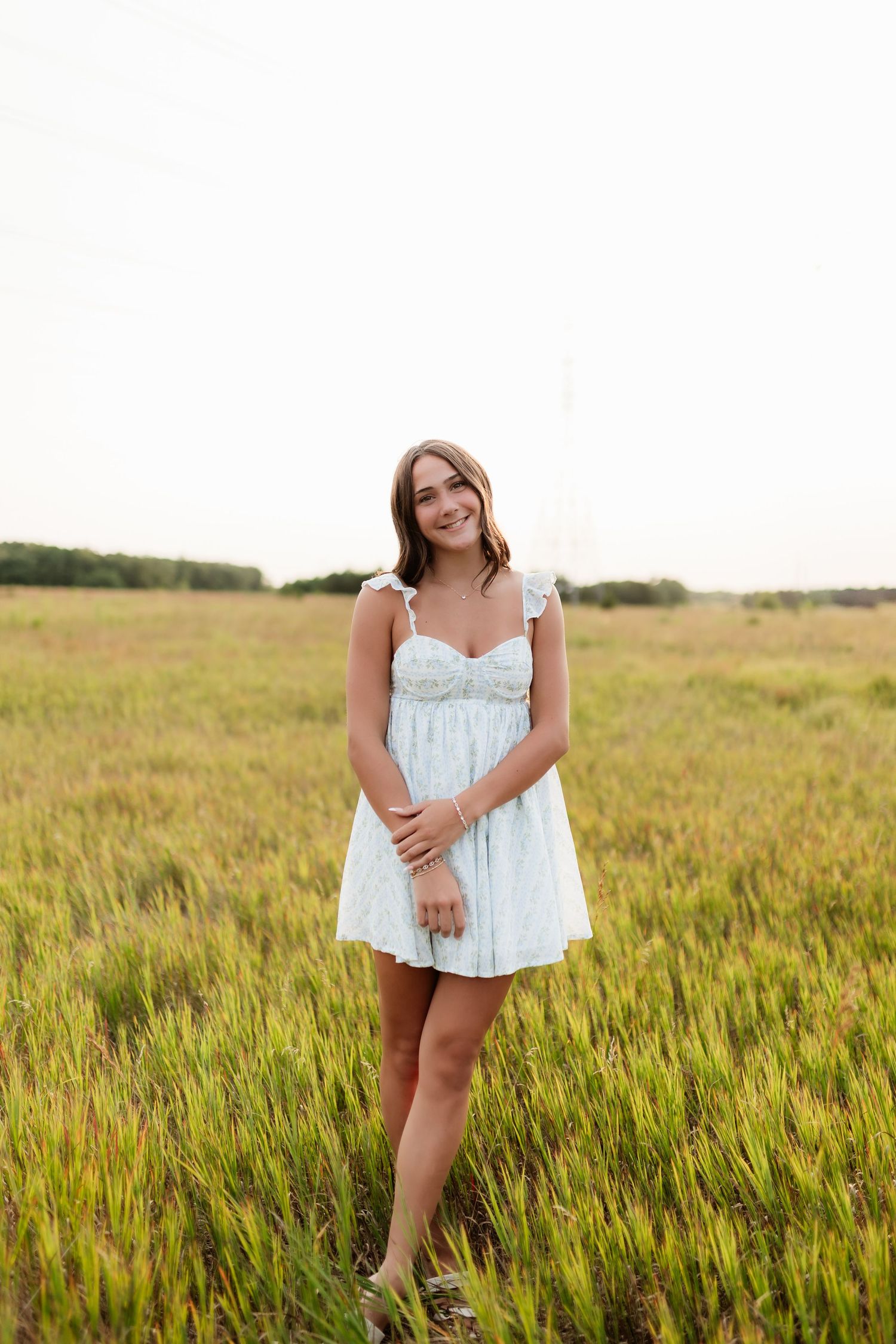 Young woman in a light blue sundress smiling in a green field on a cloudy day.