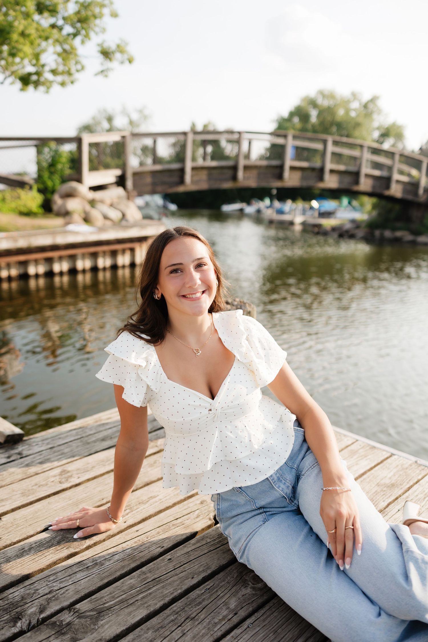 Young woman in white ruffled top and jeans sitting on wooden dock by a serene waterway with bridge backdrop.