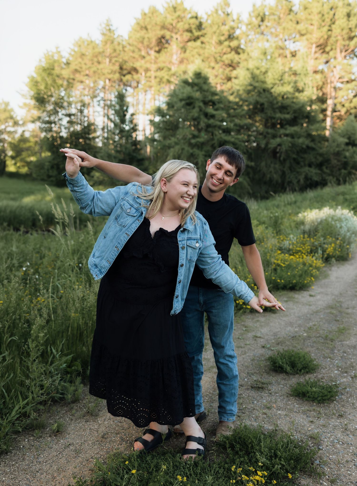 A smiling couple walks on a dirt path surrounded by green fields and trees, the woman pointing and wearing a denim jacket.