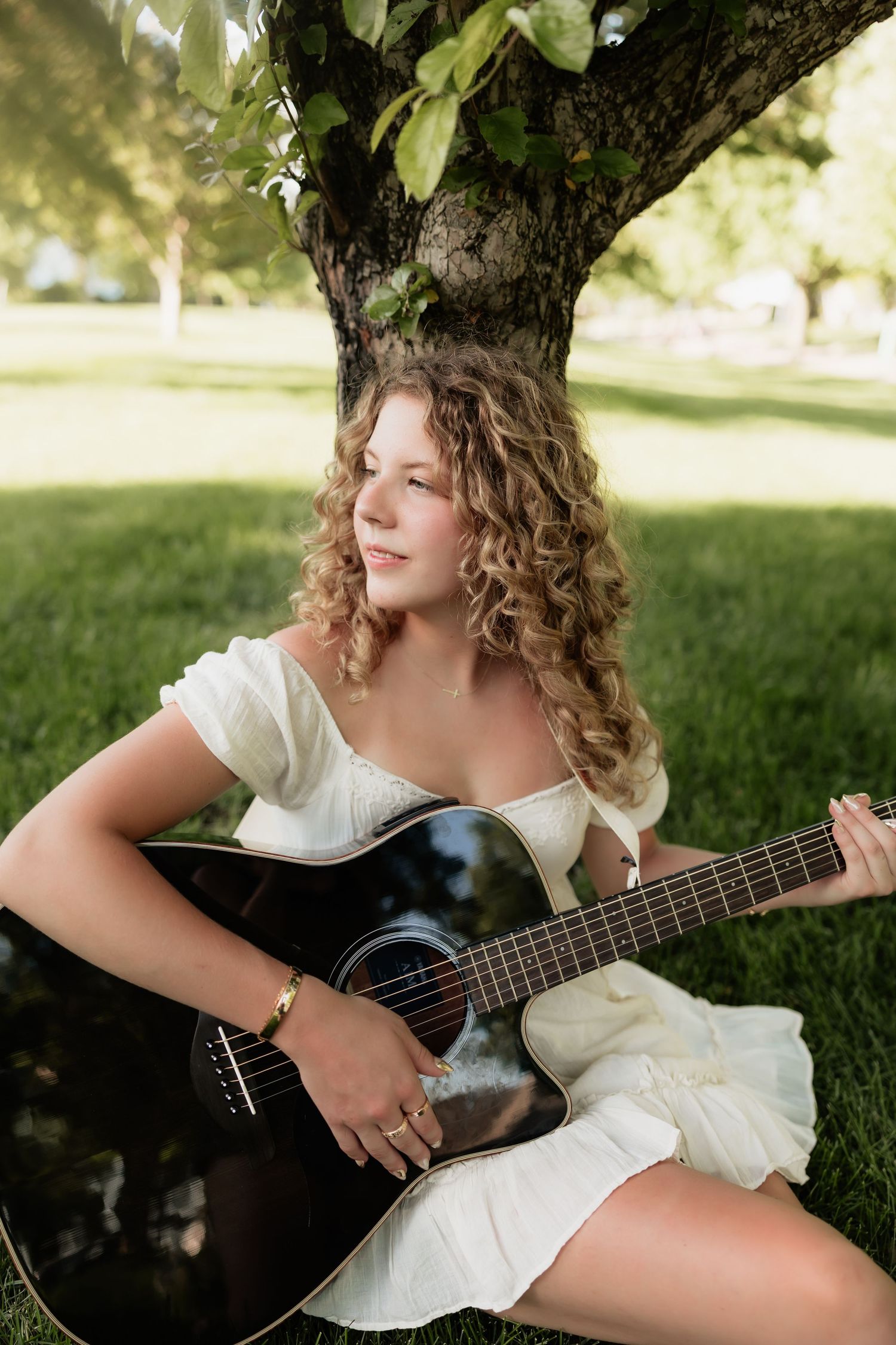 Smiling young woman with curly hair holding black acoustic guitar while sitting under a tree in a white sundress.