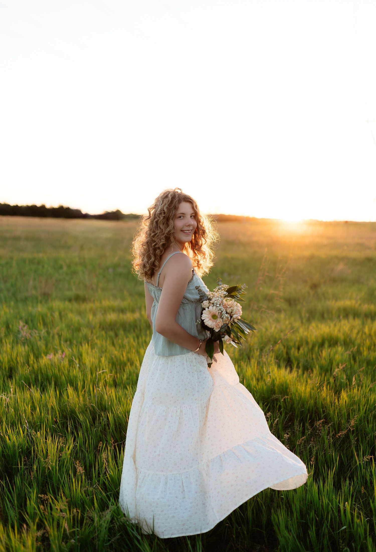 Young woman with curly hair holding a bouquet in a green field during a vibrant golden sunset.
