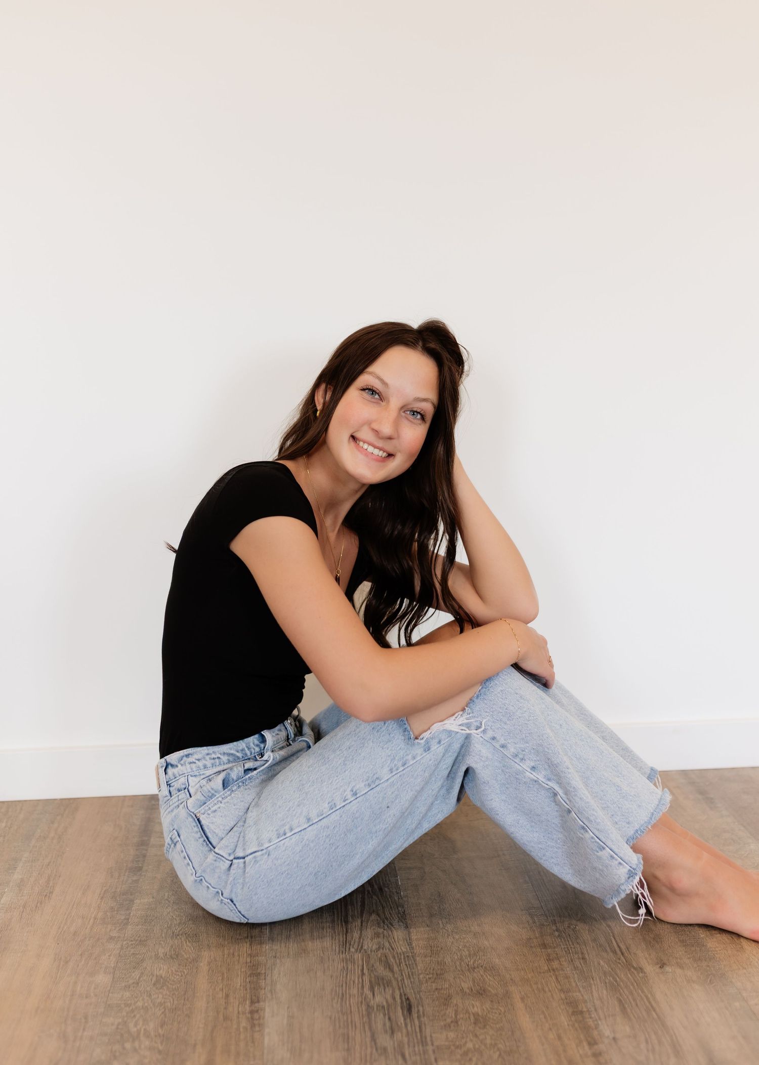 Young woman sitting on floor smiling, wearing black top and light wash jeans.