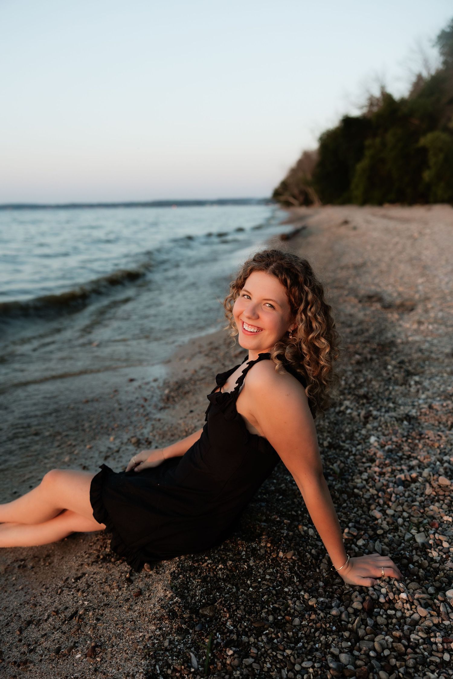 Smiling woman with curly hair in black dress sitting on pebble beach at sunset with calm water behind her.