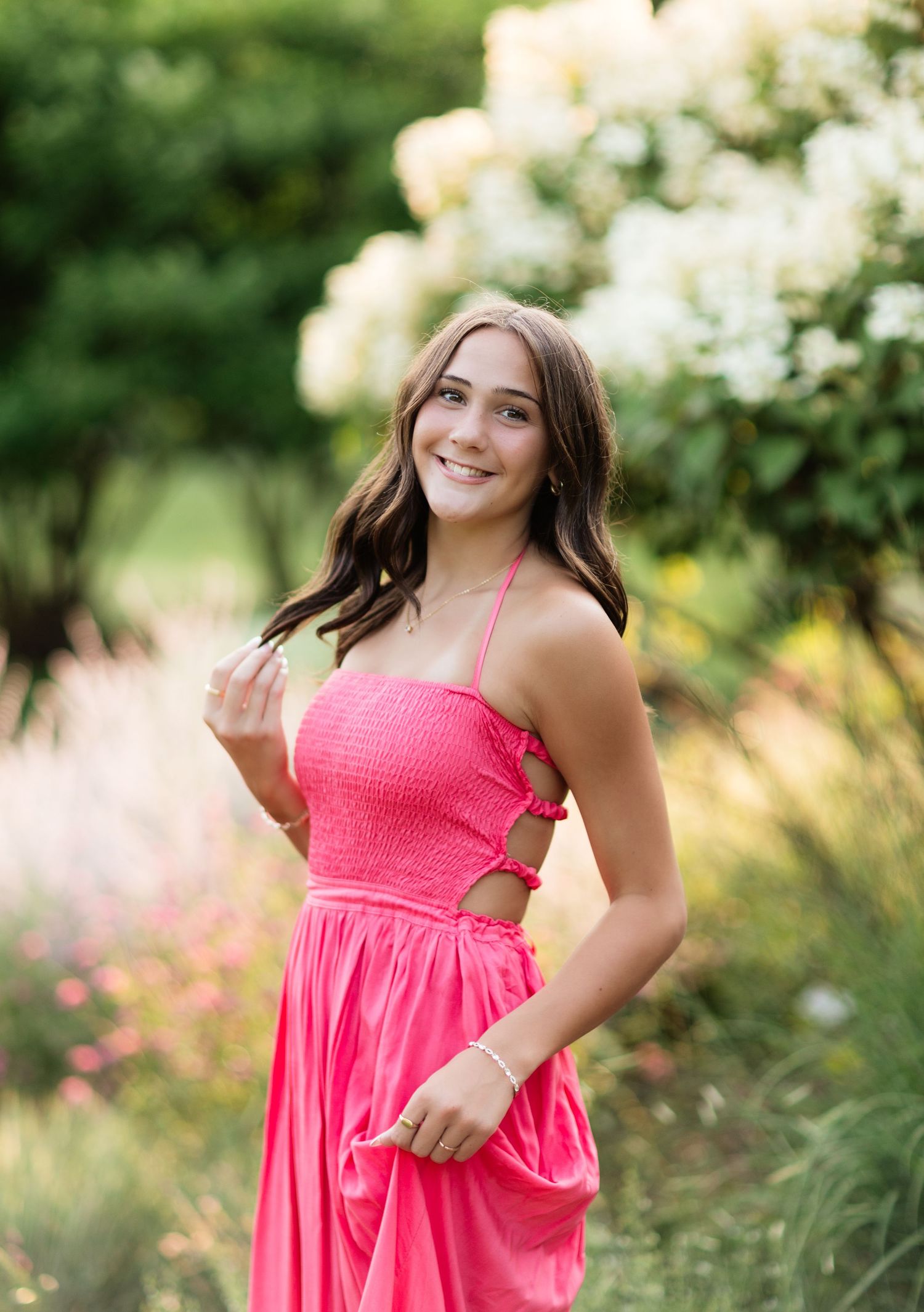Brunette teen smiling in a bright pink smocked halter dress surrounded by blooming garden flowers.