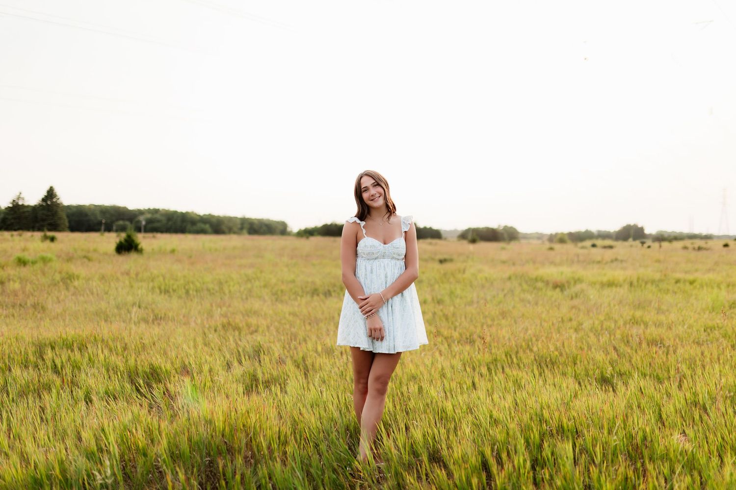 Teen girl in white sundress standing in open green field at golden hour.