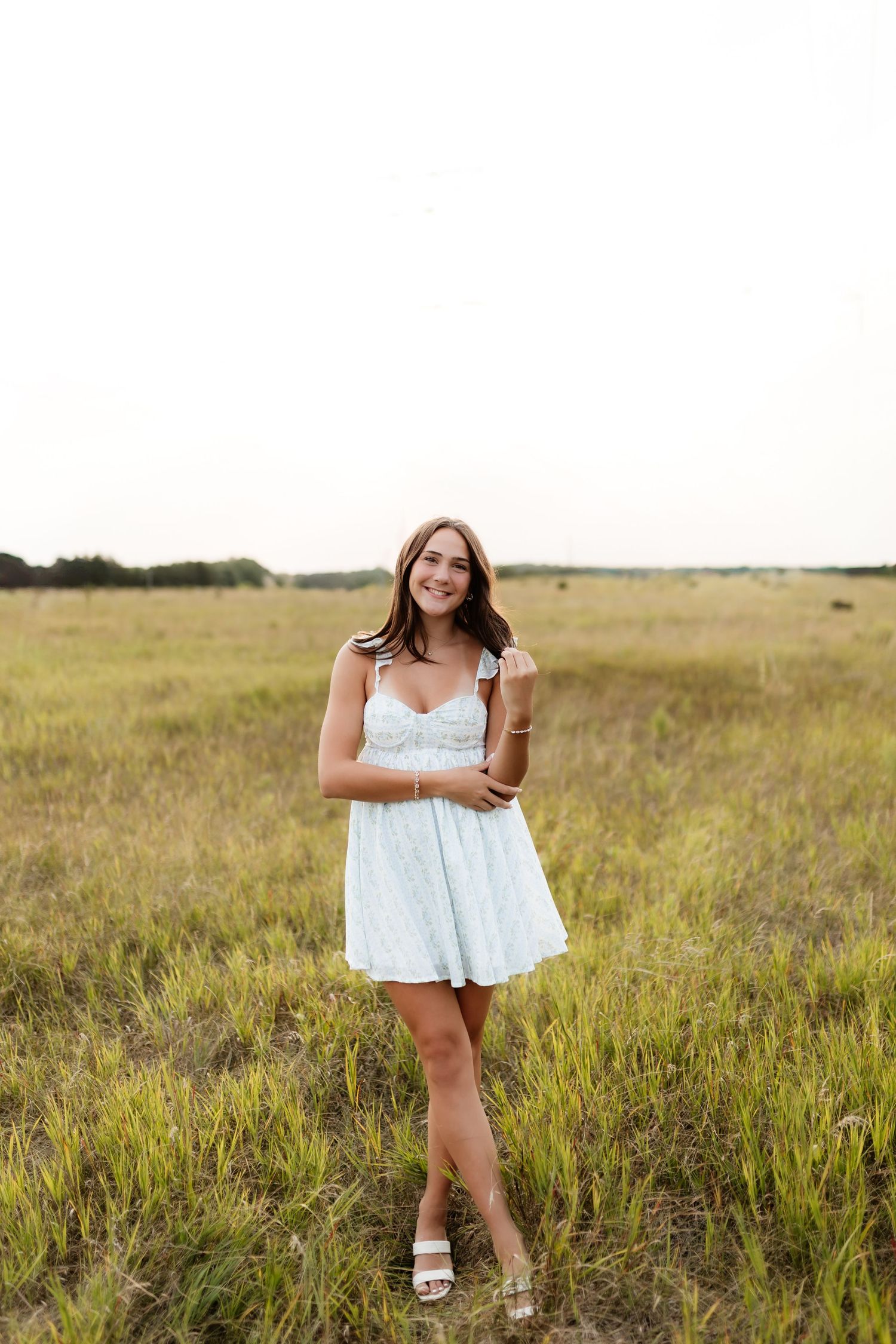 Young woman in white floral sundress smiling and posing in a wide open grassy meadow.