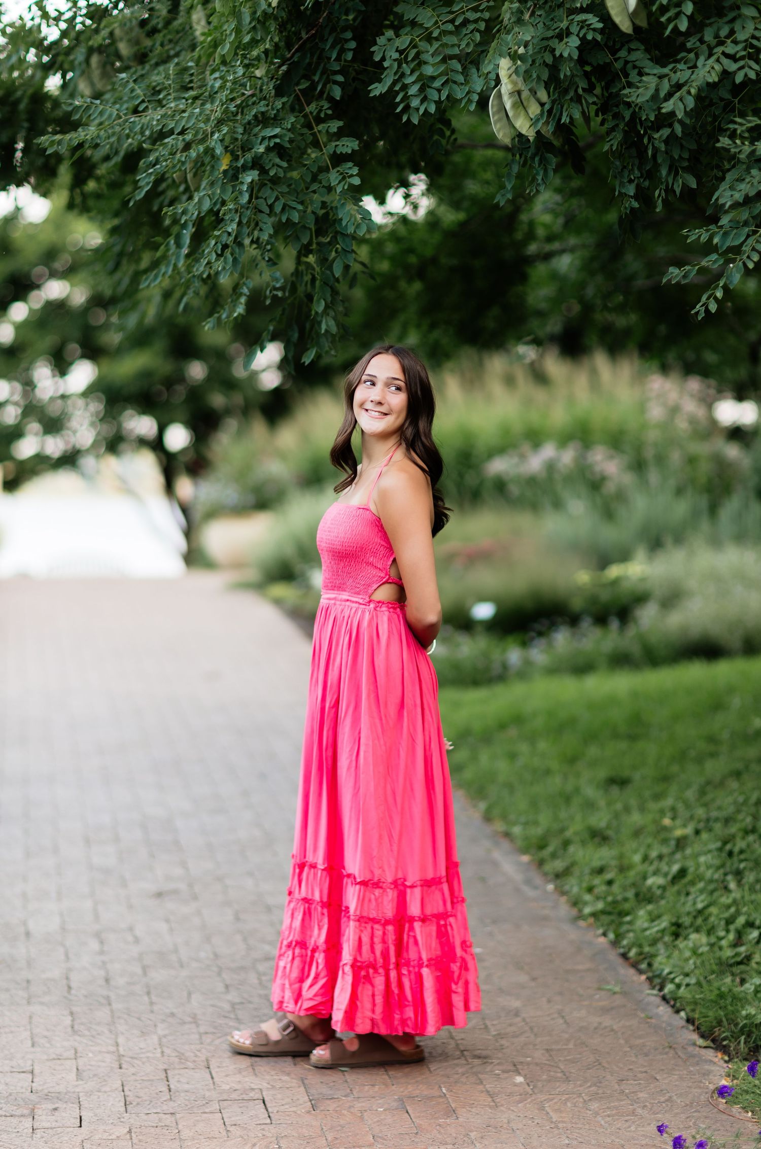 Young woman in bright pink maxi dress smiling in lush garden pathway.