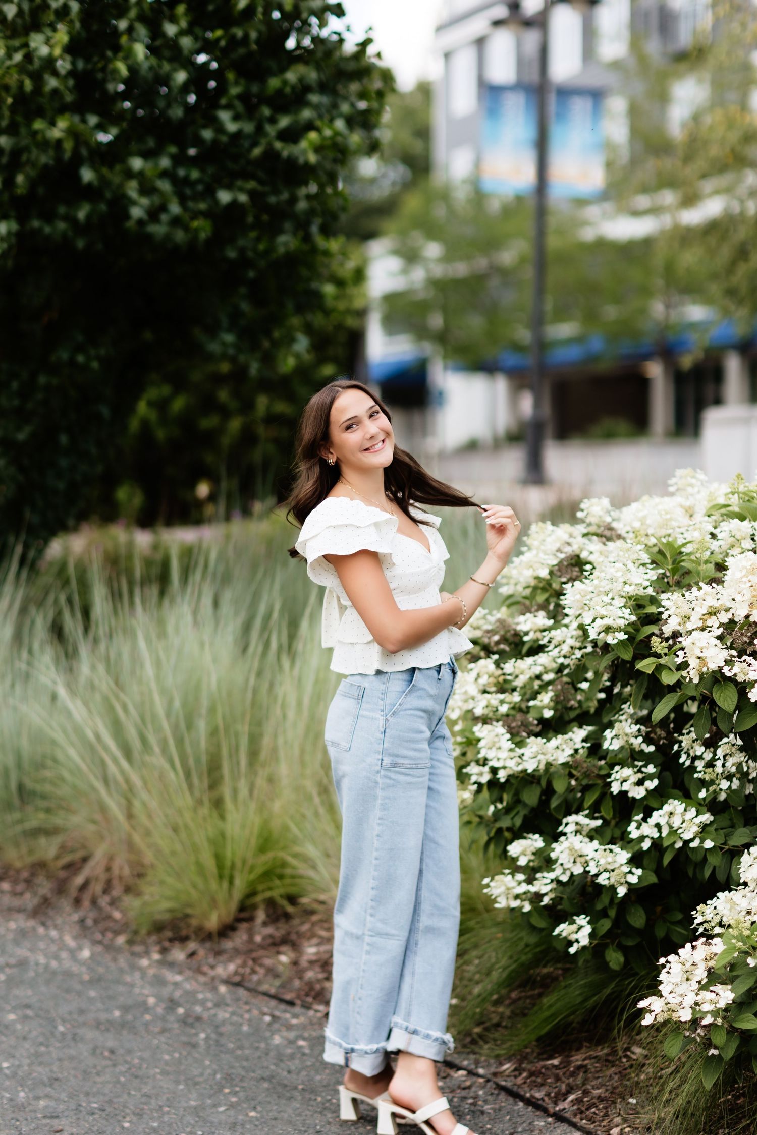 Smiling brunette woman in white top and blue jeans posing near white hydrangea bushes outdoors.