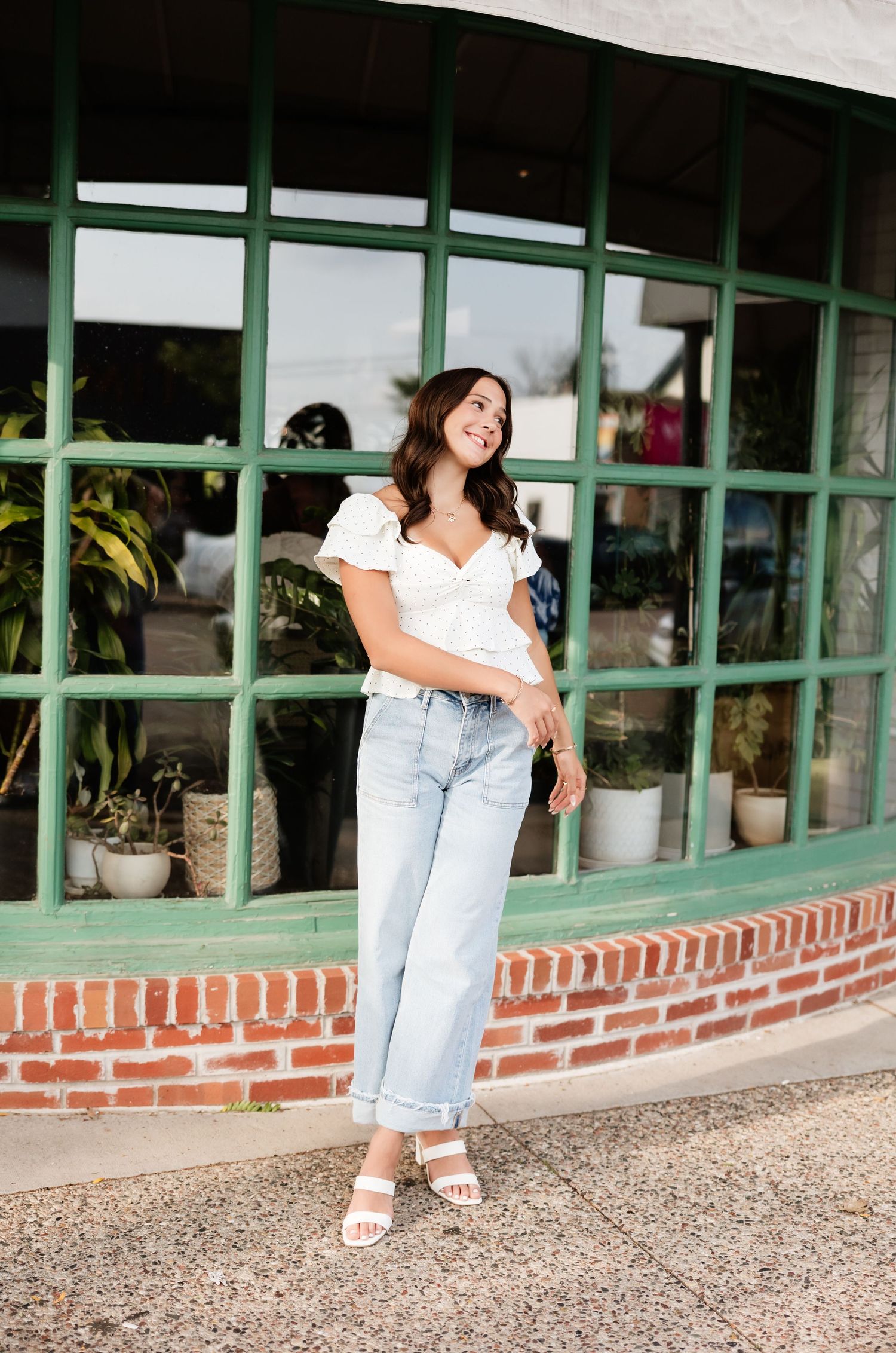 Smiling young woman in white crop top and cuffed light jeans standing before a green-framed shop window.