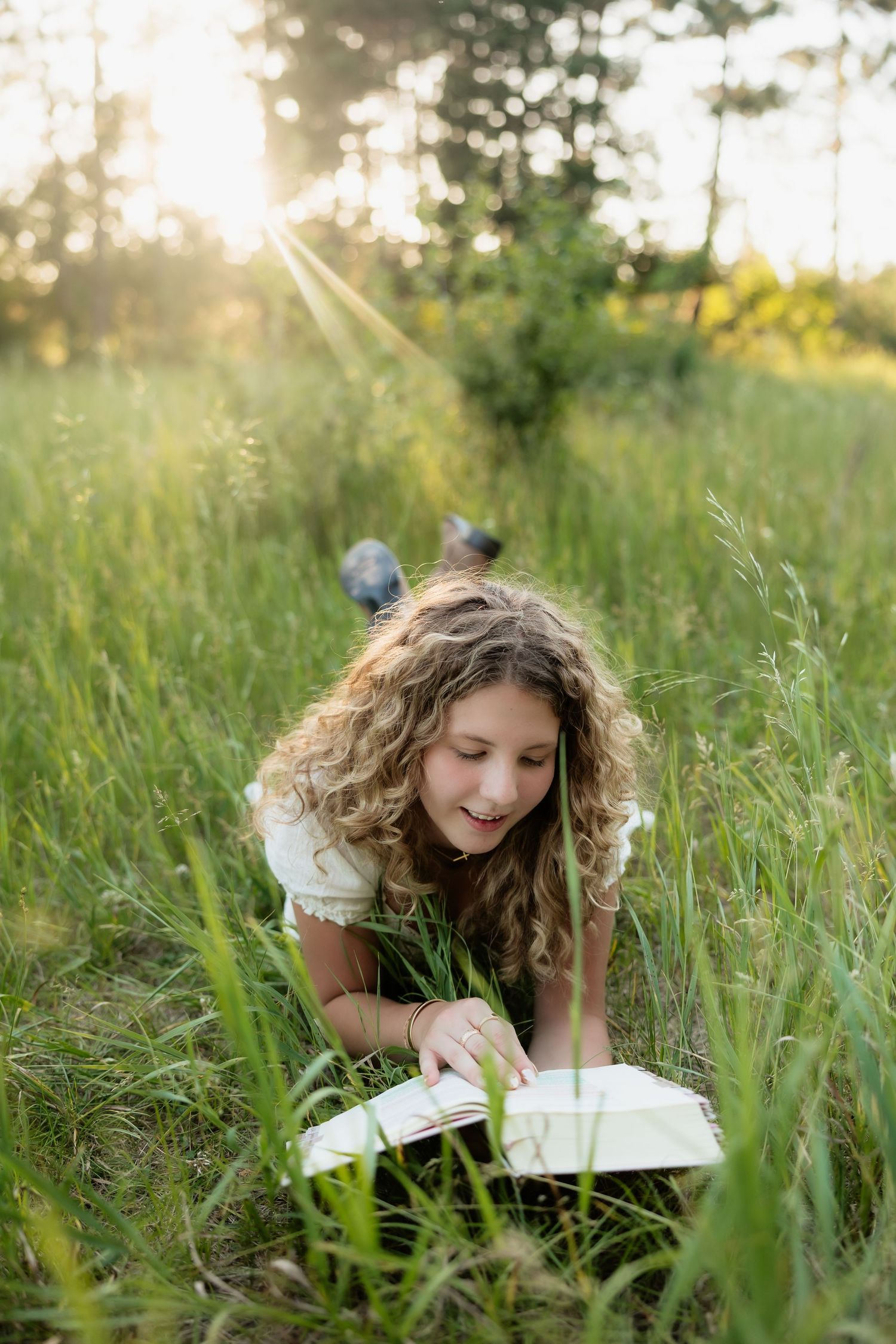 Curly haired girl lying in tall green grass reading book in golden sunlight.
