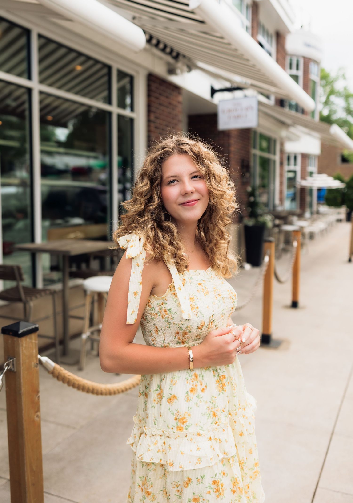 Young woman with curly hair smiling in a floral yellow dress on a sunny outdoor shopping street.