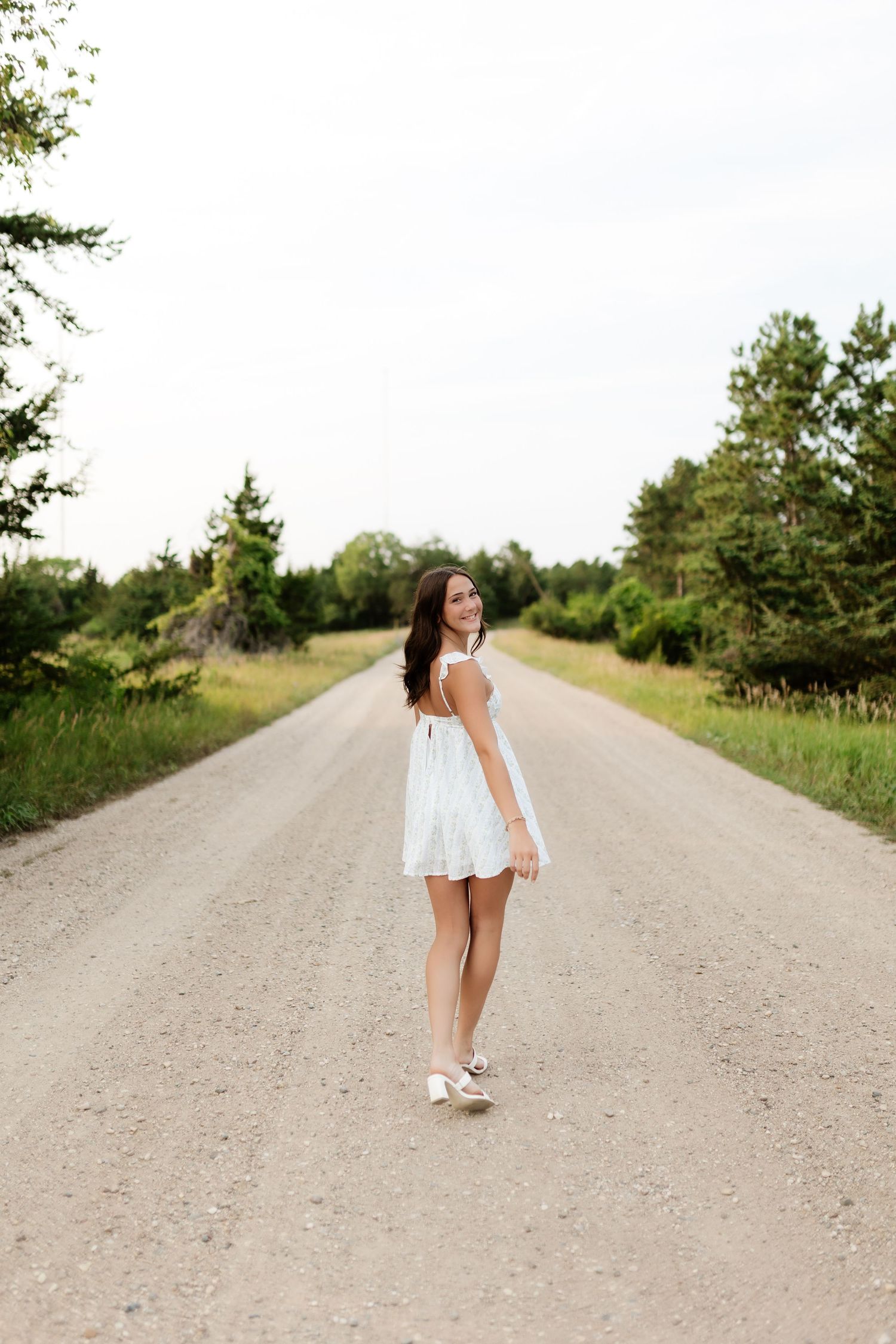 Young woman in white sundress looking back over shoulder while standing on scenic gravel road.