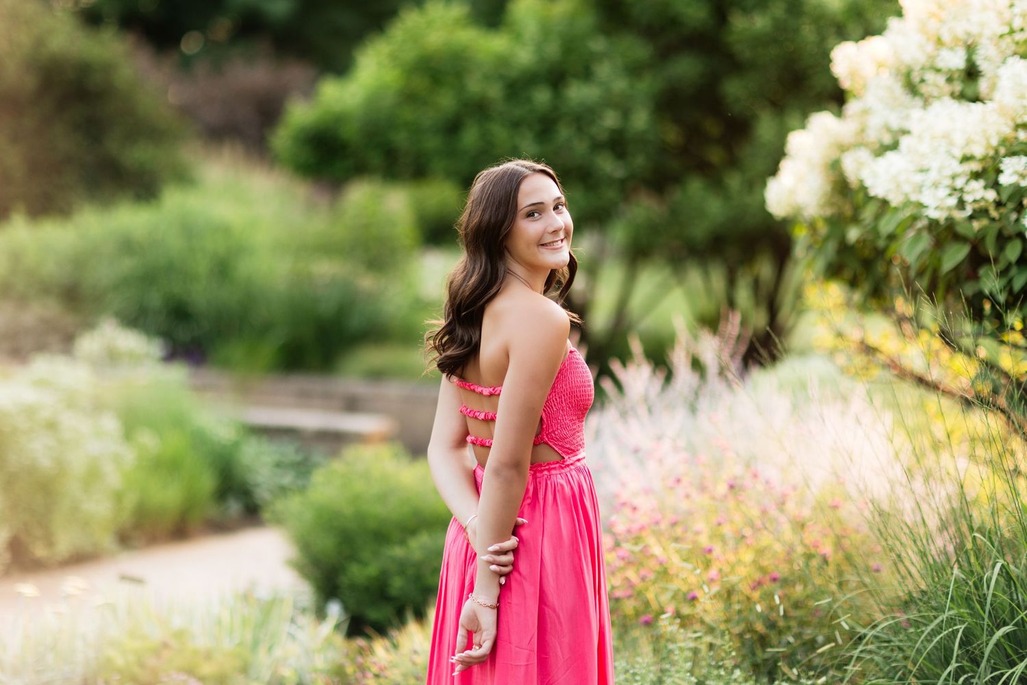 Young woman in pink strapless maxi dress standing in a lush garden, glancing back over shoulder.