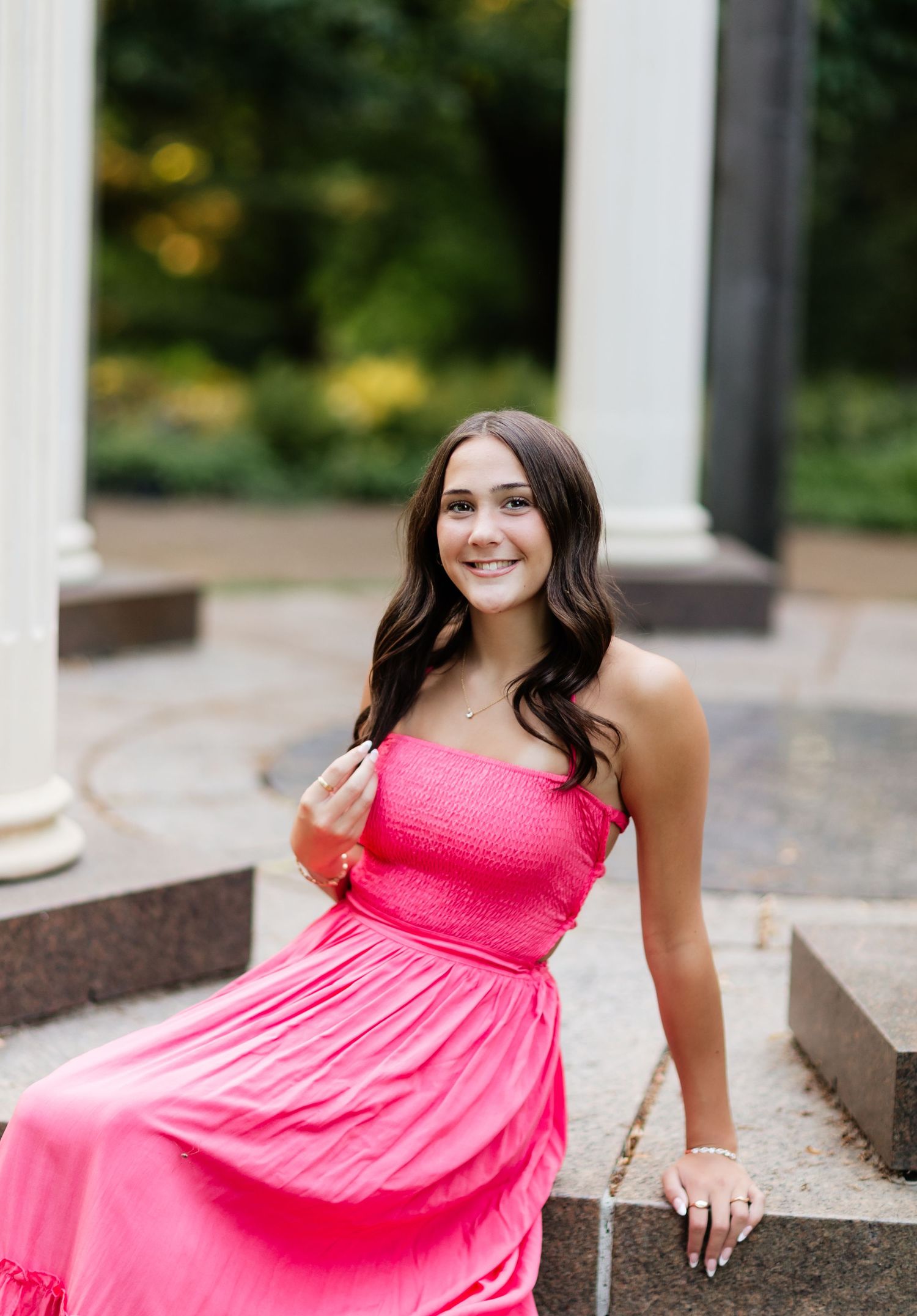 Smiling teen girl in pink strapless maxi dress seated near white columns in an outdoor garden setting.