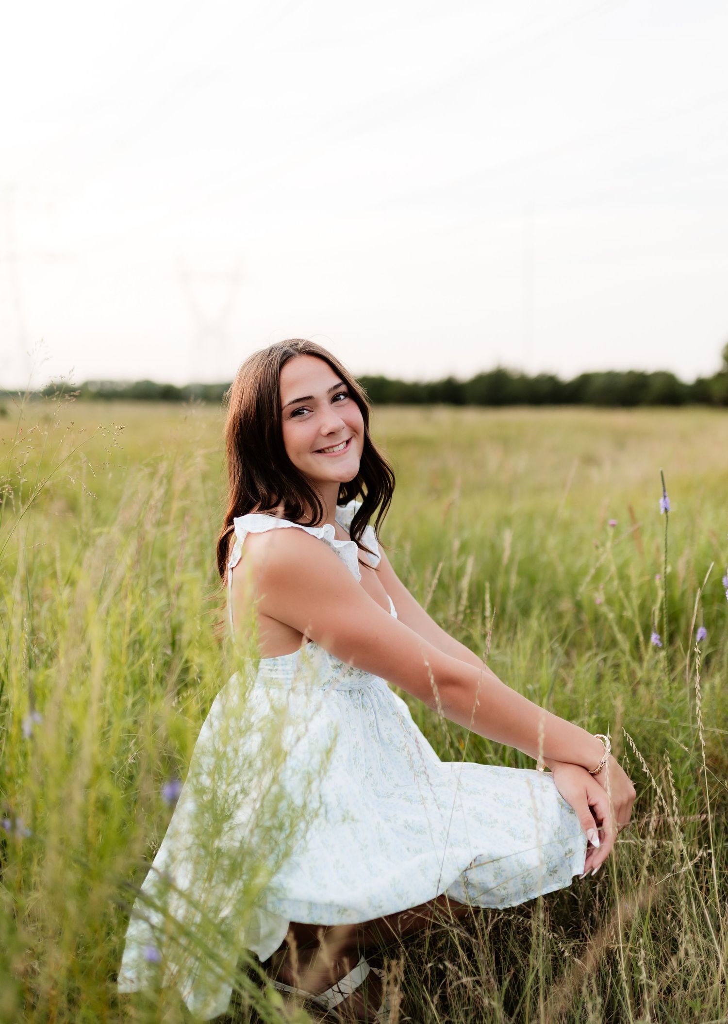 Smiling young woman in white floral dress sitting in tall grass in a sunlit open meadow.
