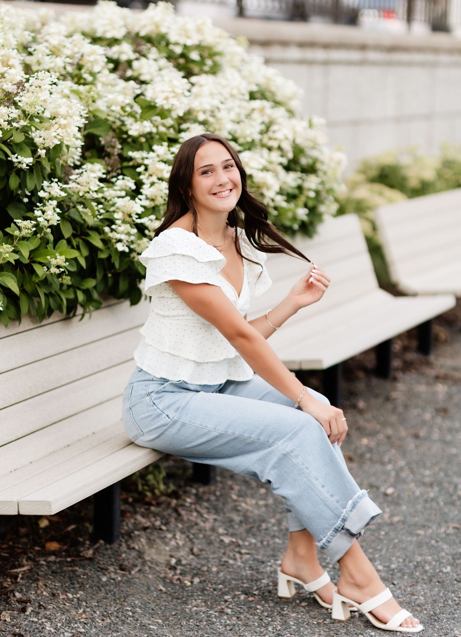 Young woman in white top and cuffed jeans laughing on bench surrounded by white hydrangea blooms.