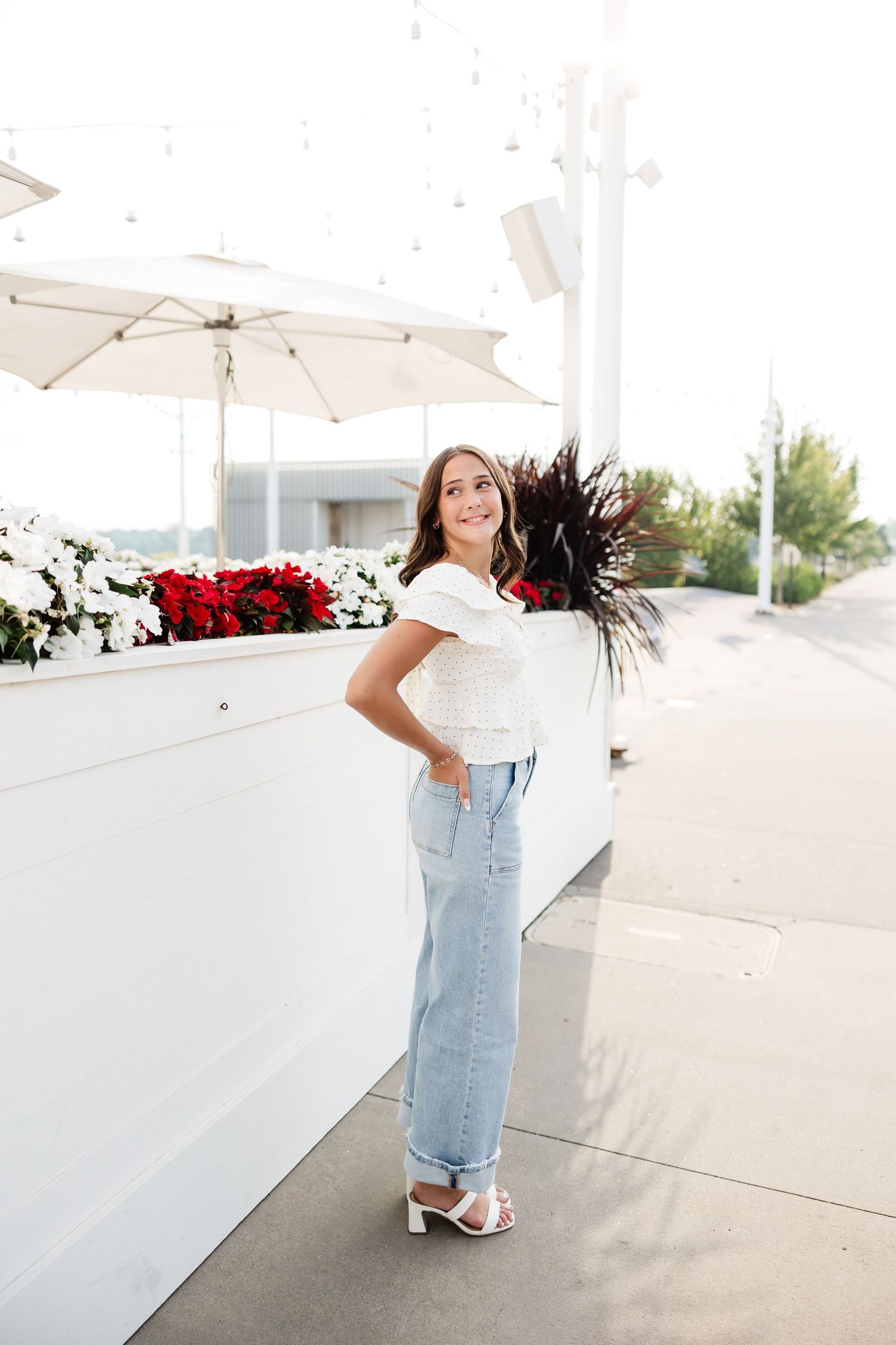 Young woman in white top and light jeans leaning against a white wall with red and white flowers.