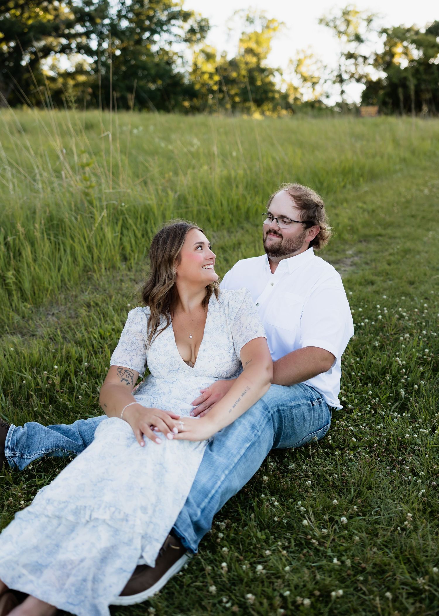 Couple in white outfits sitting together in a lush green field, smiling and holding hands during golden hour.