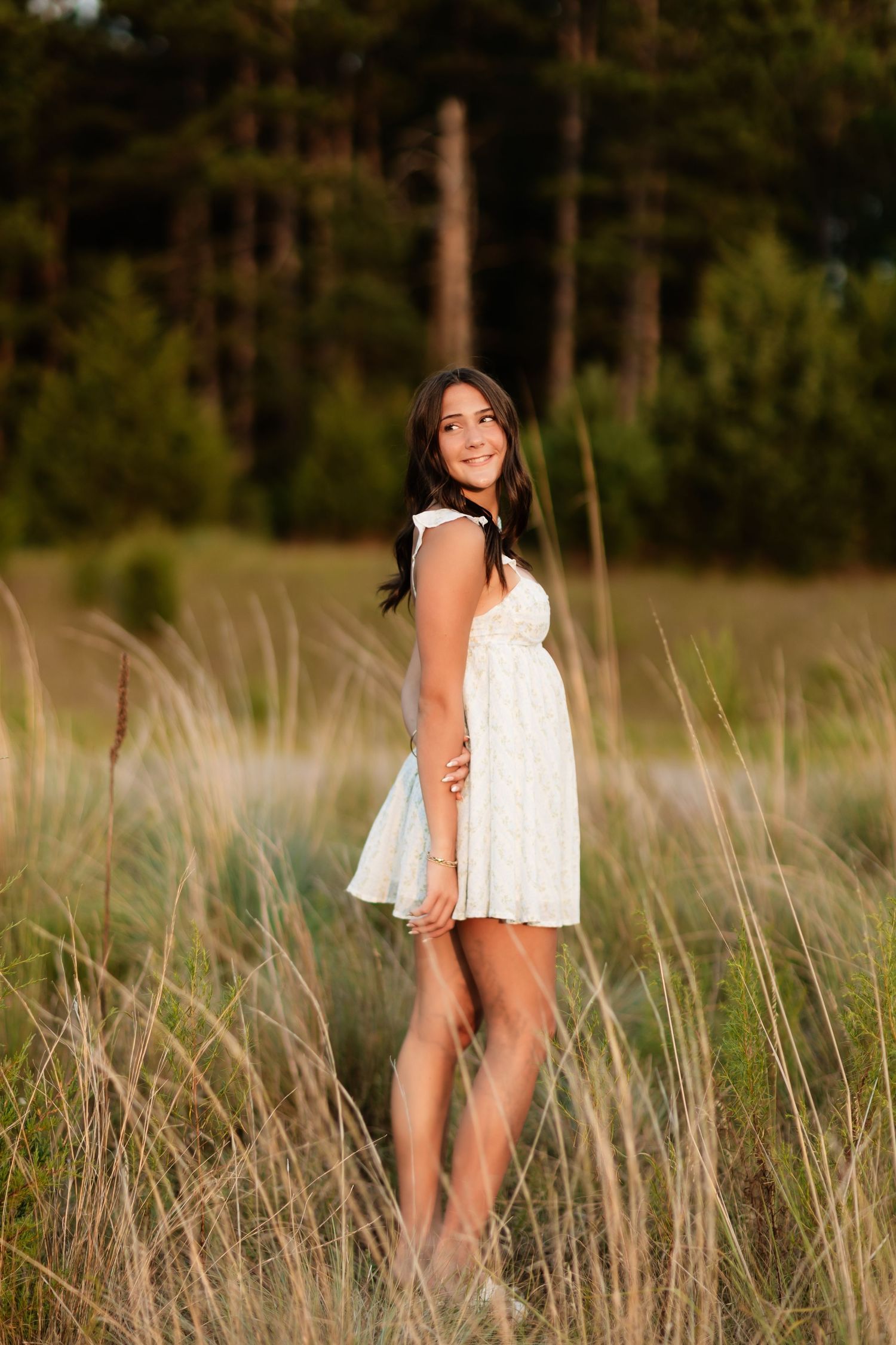Young woman in white dress posing in tall grass with pine trees background.
