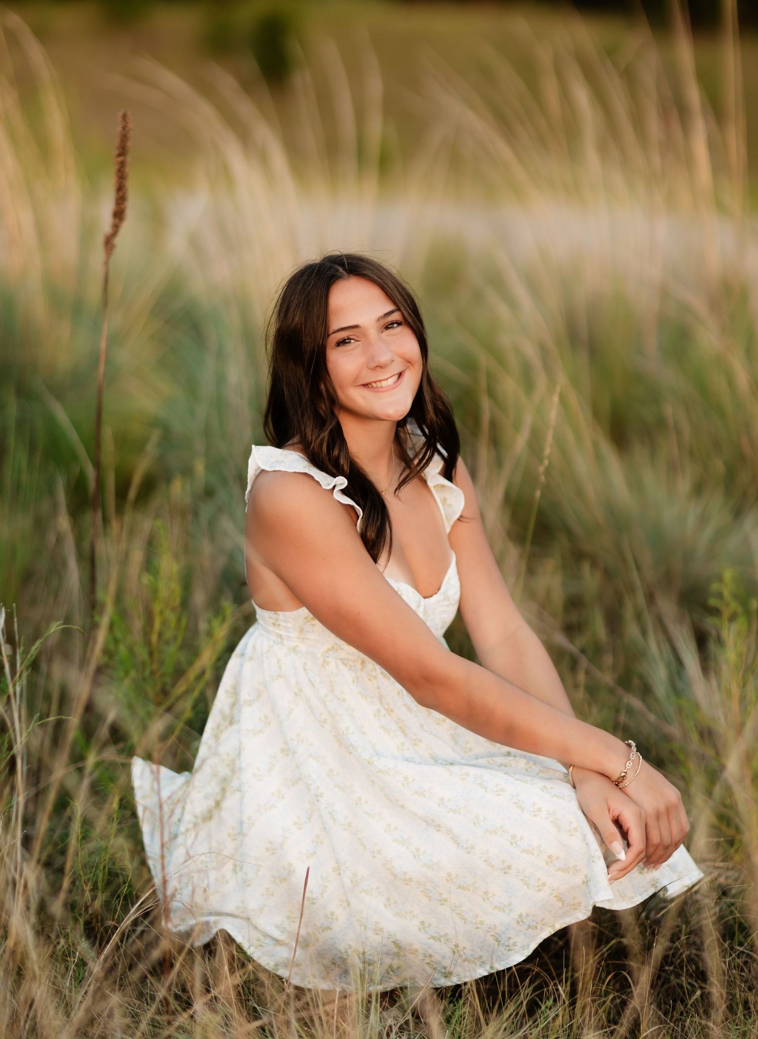 Young woman in white floral dress sitting gracefully among tall golden grasses in a field.