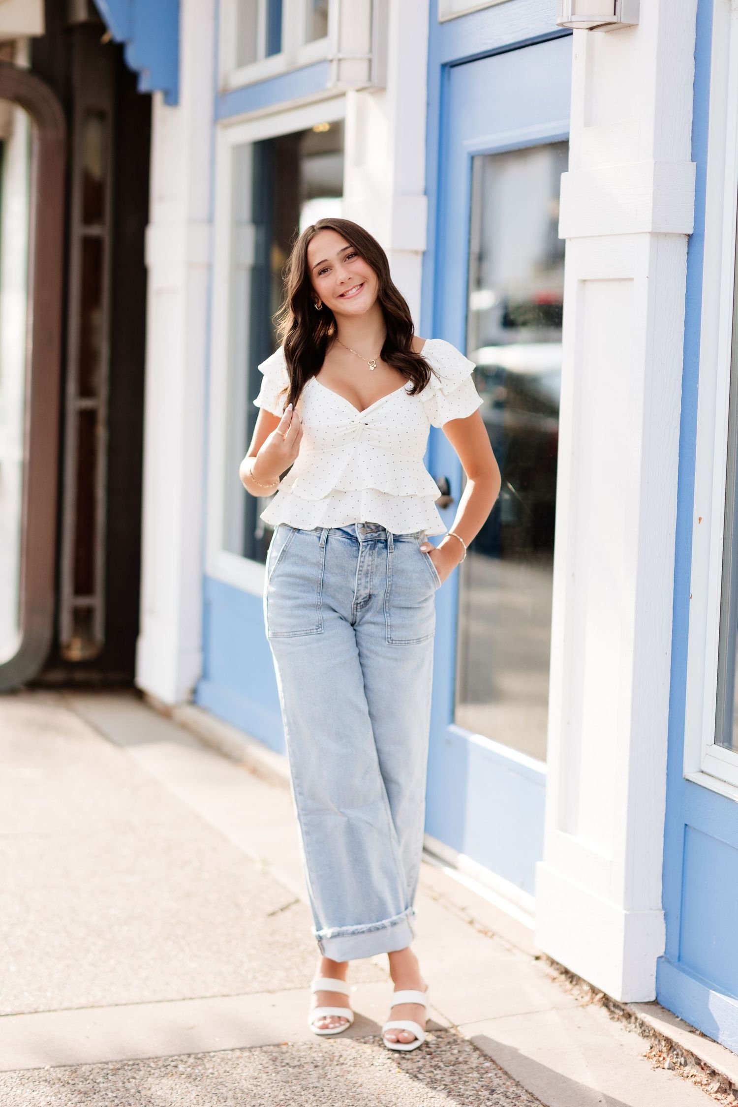 Smiling young woman in white eyelet top and light jeans posing outside blue and white storefront.