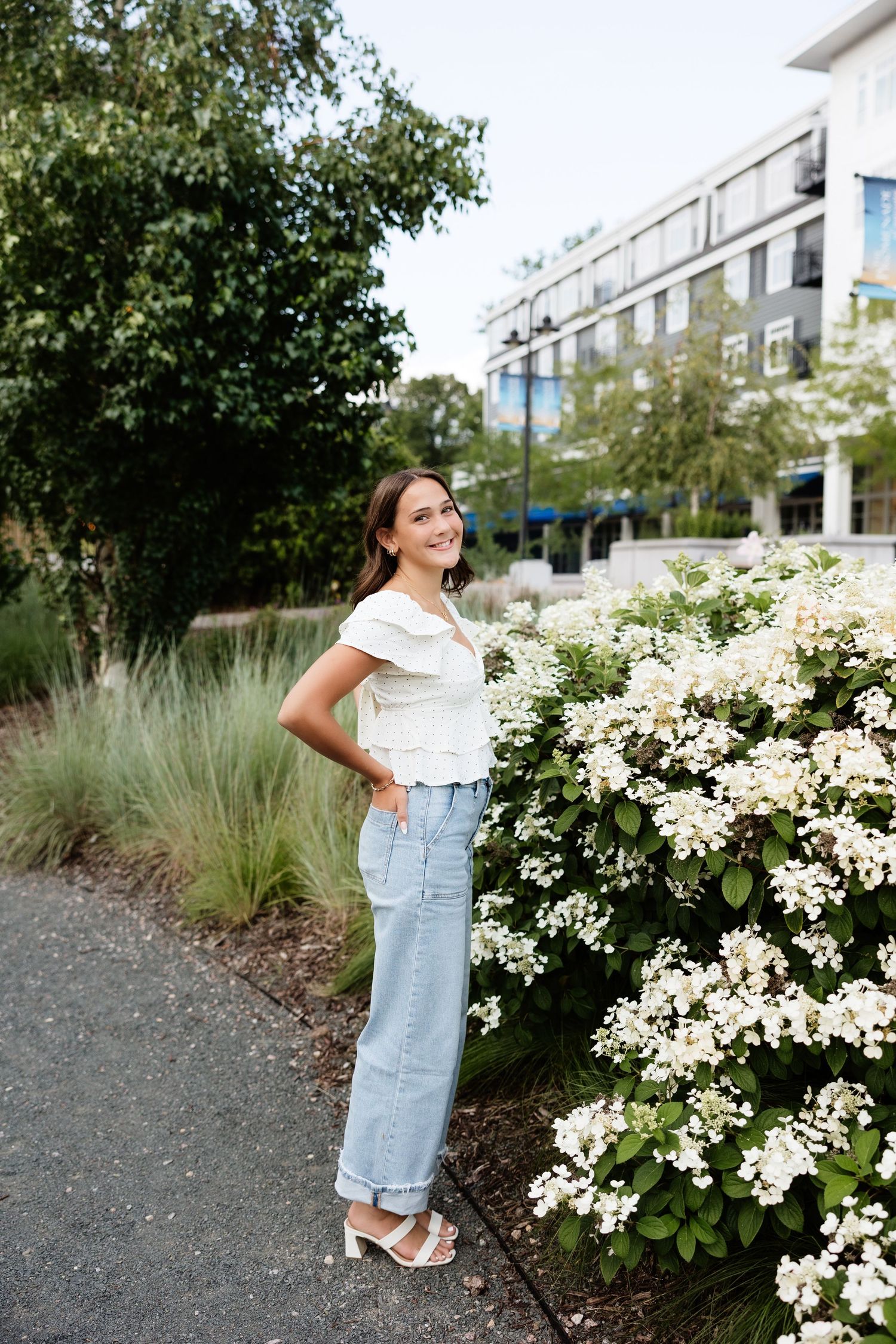 Young woman in white top and jeans smiling beside white flowering bush in urban garden setting.