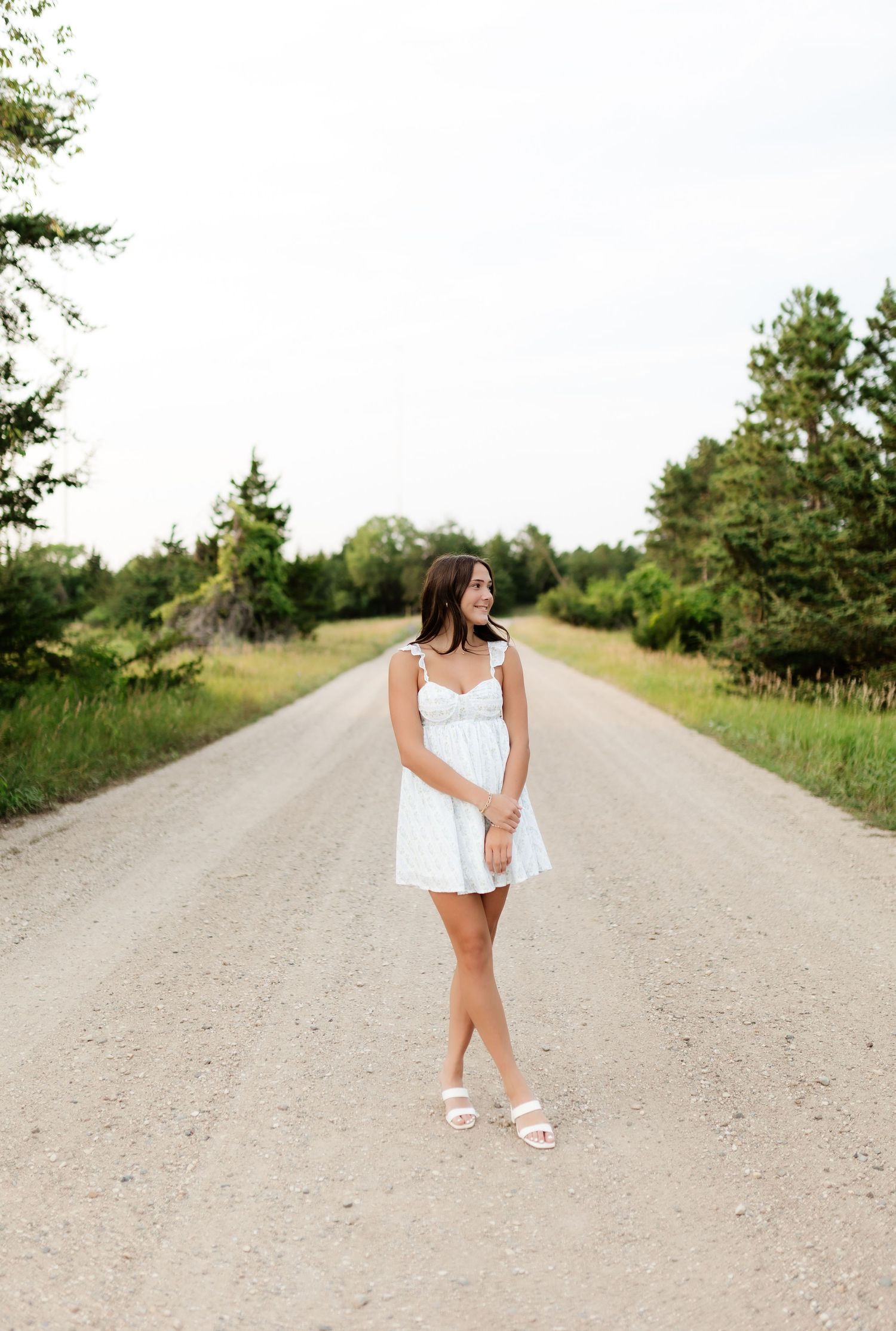 Young woman in white dress walking on a scenic gravel road flanked by green trees.
