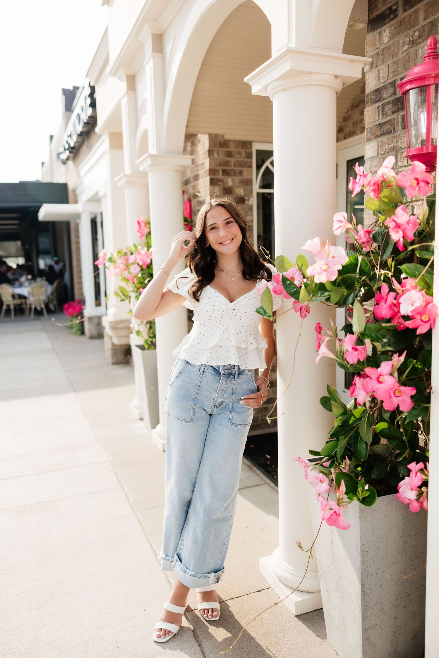 Brunette woman in white peplum top and jeans posing beside pink bougainvillea flowers on patio.