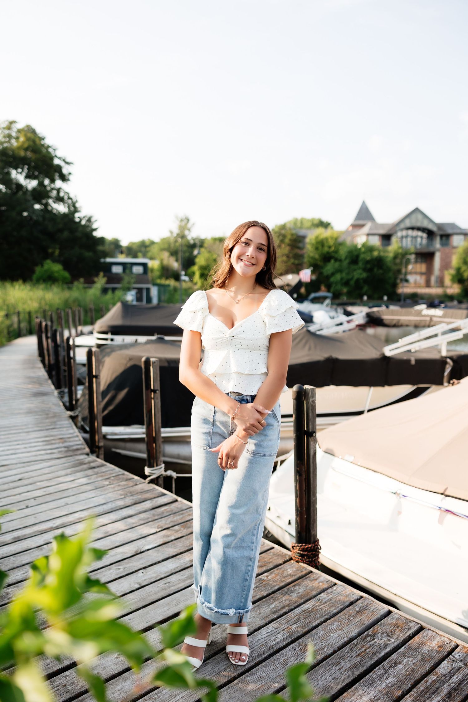 Smiling young woman in white top and jeans standing on a wooden dock with boats in background.