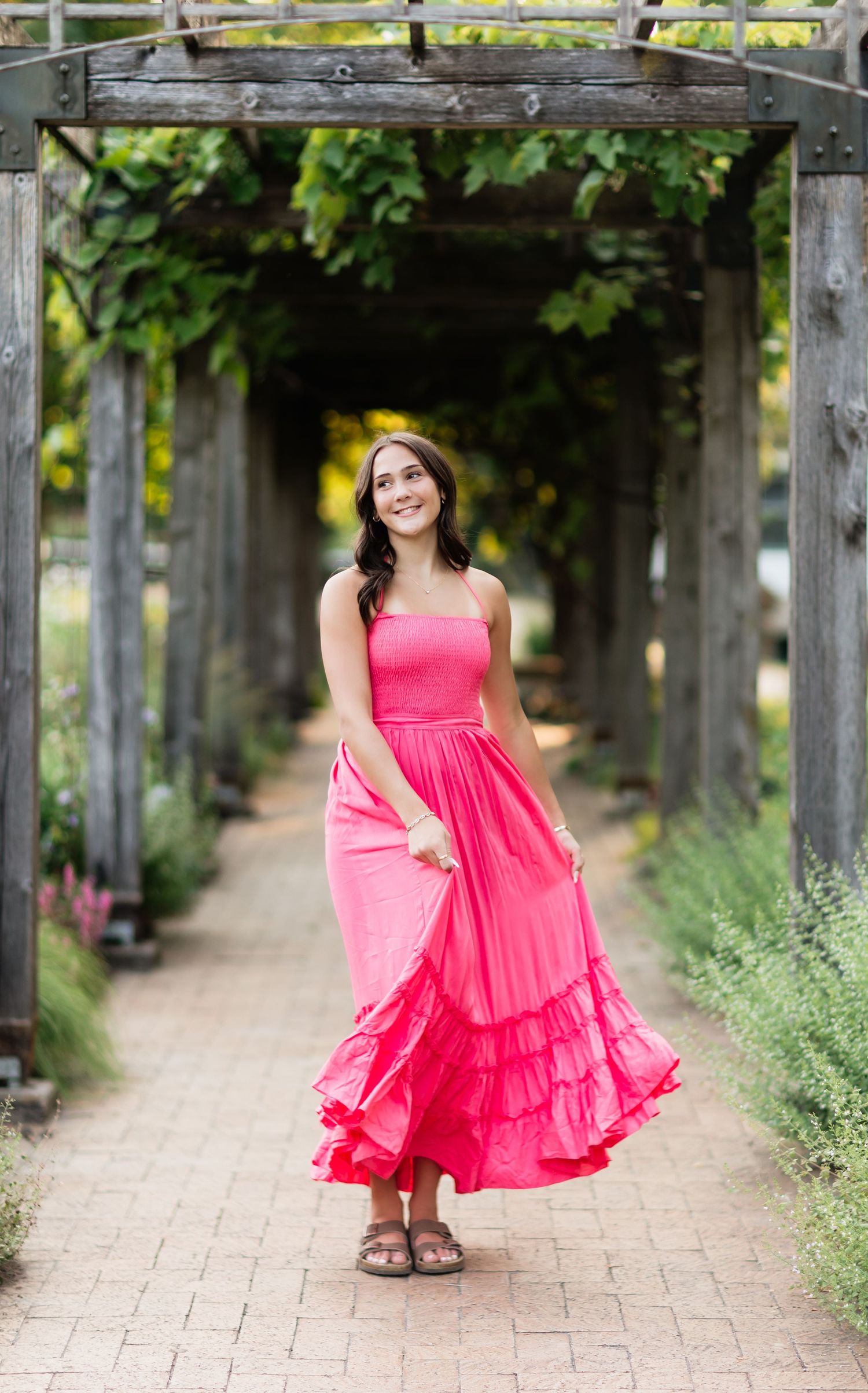 Young woman in hot pink strapless ruffle maxi dress twirling under rustic garden pergola pathway.