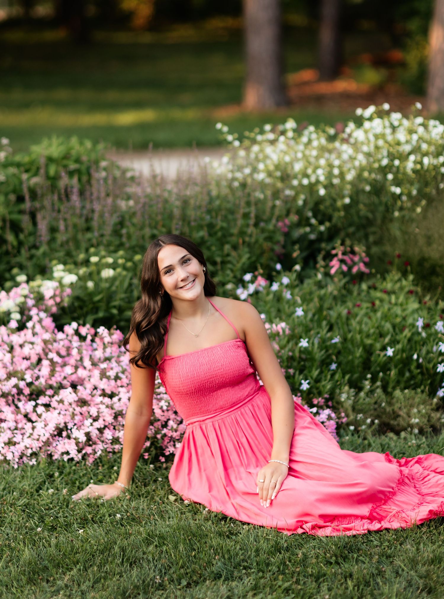 Smiling young woman in pink maxi dress sitting on grass beside a colorful flower garden.