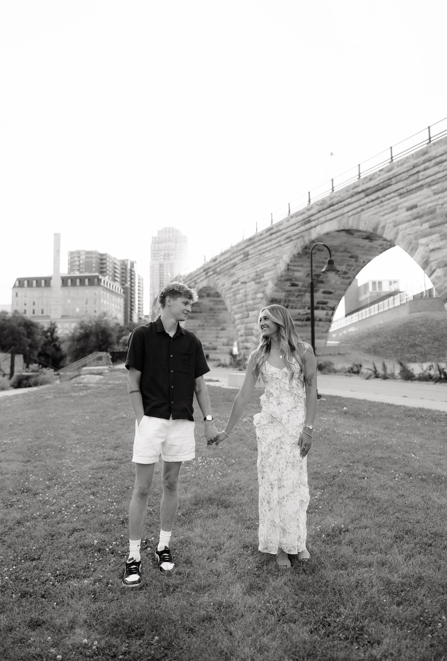 Black and white photo of a couple holding hands on a grassy lawn near a stone bridge.