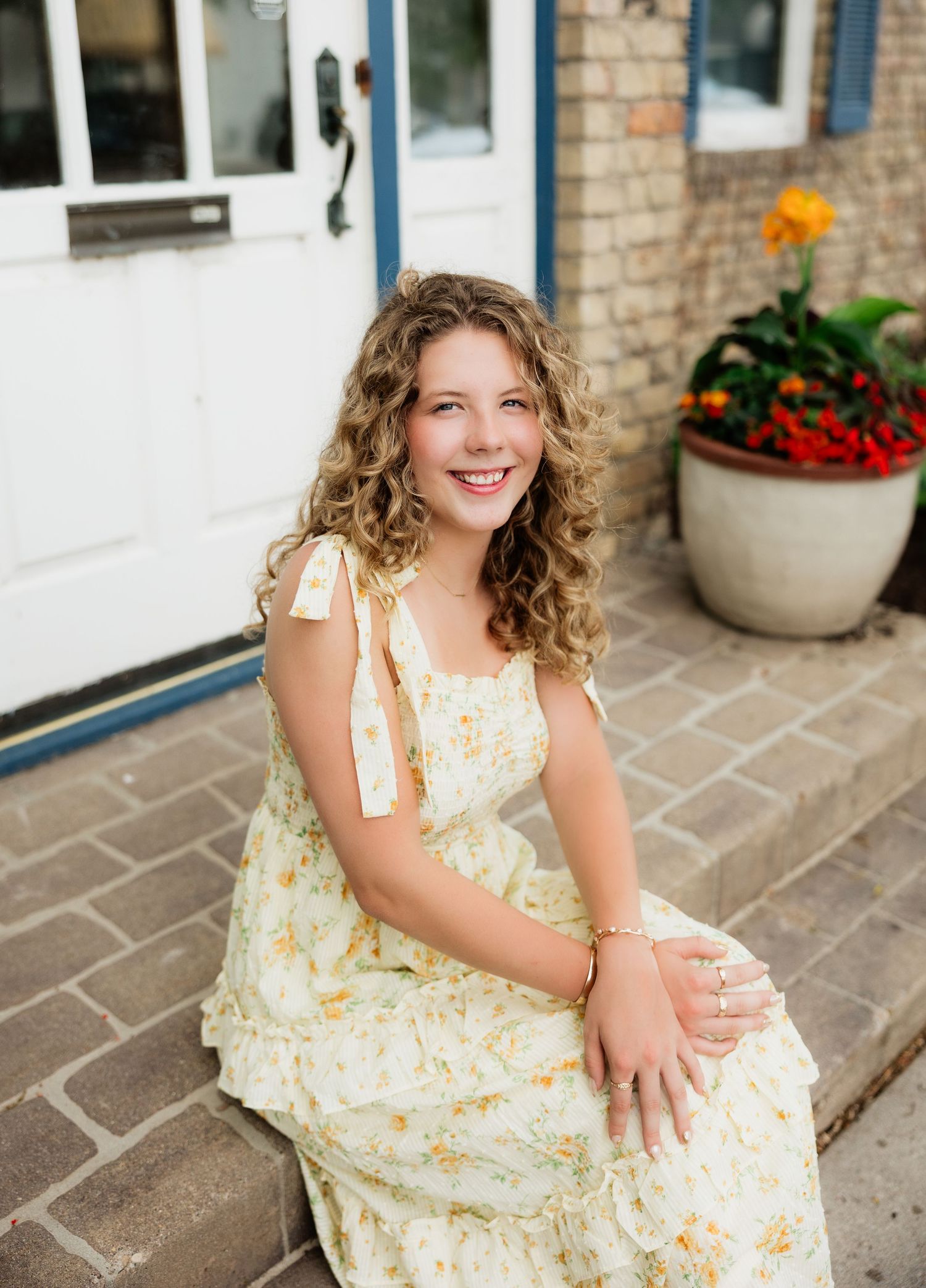 Smiling young woman with curly hair in a floral sundress sitting on brick steps with flowers.