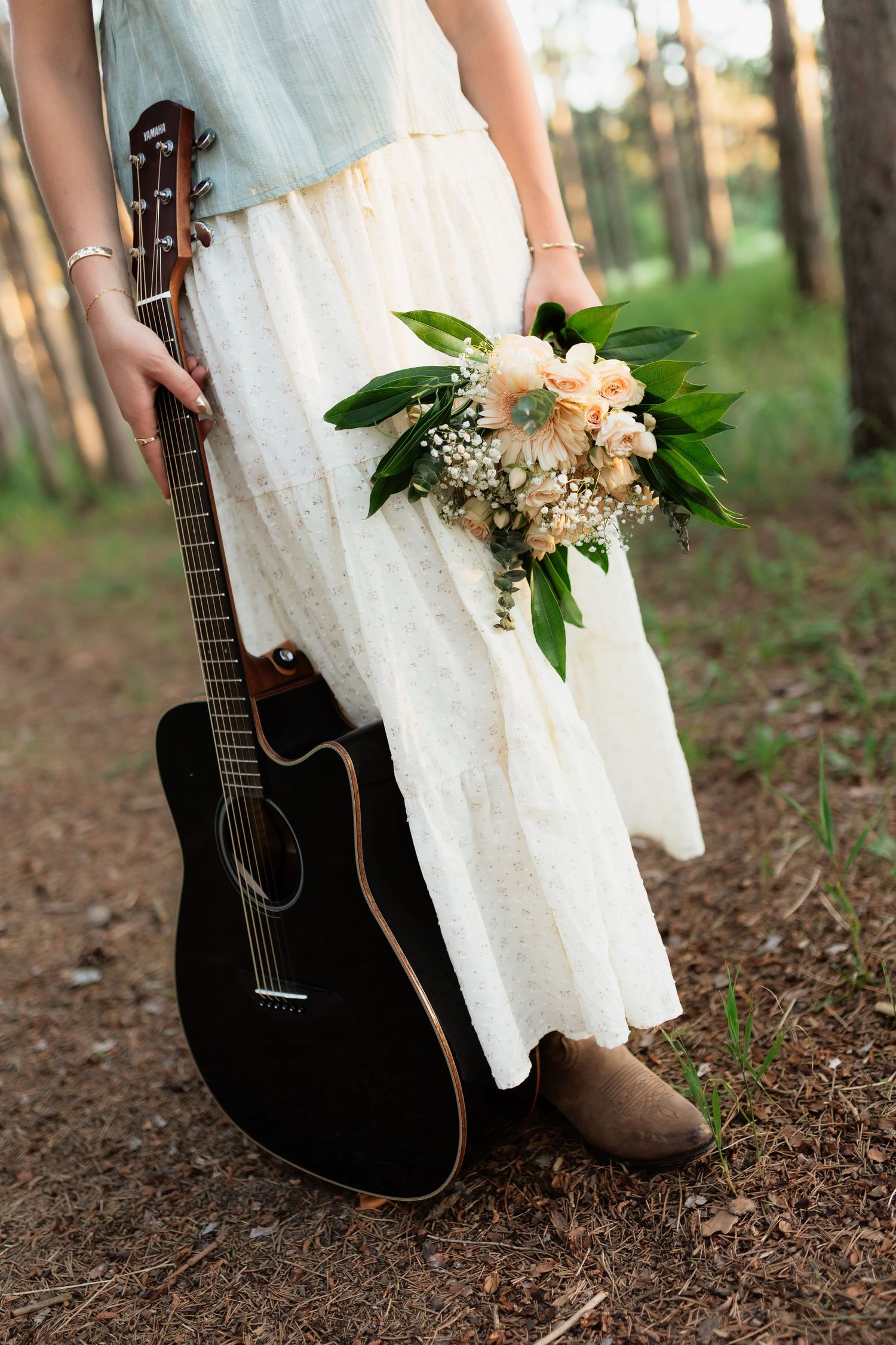 Woman in white skirt holding black acoustic guitar and floral bouquet while standing in pine forest.