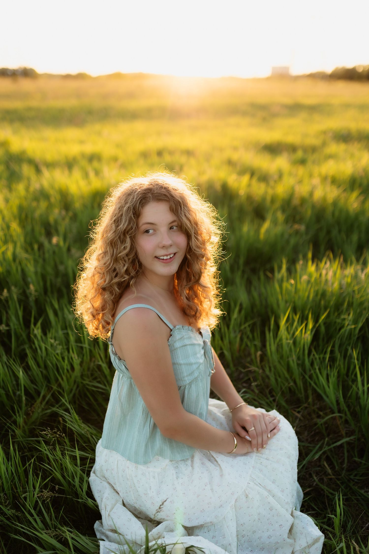 Curly-haired young woman in mint sundress sitting in green field during golden hour sunset.