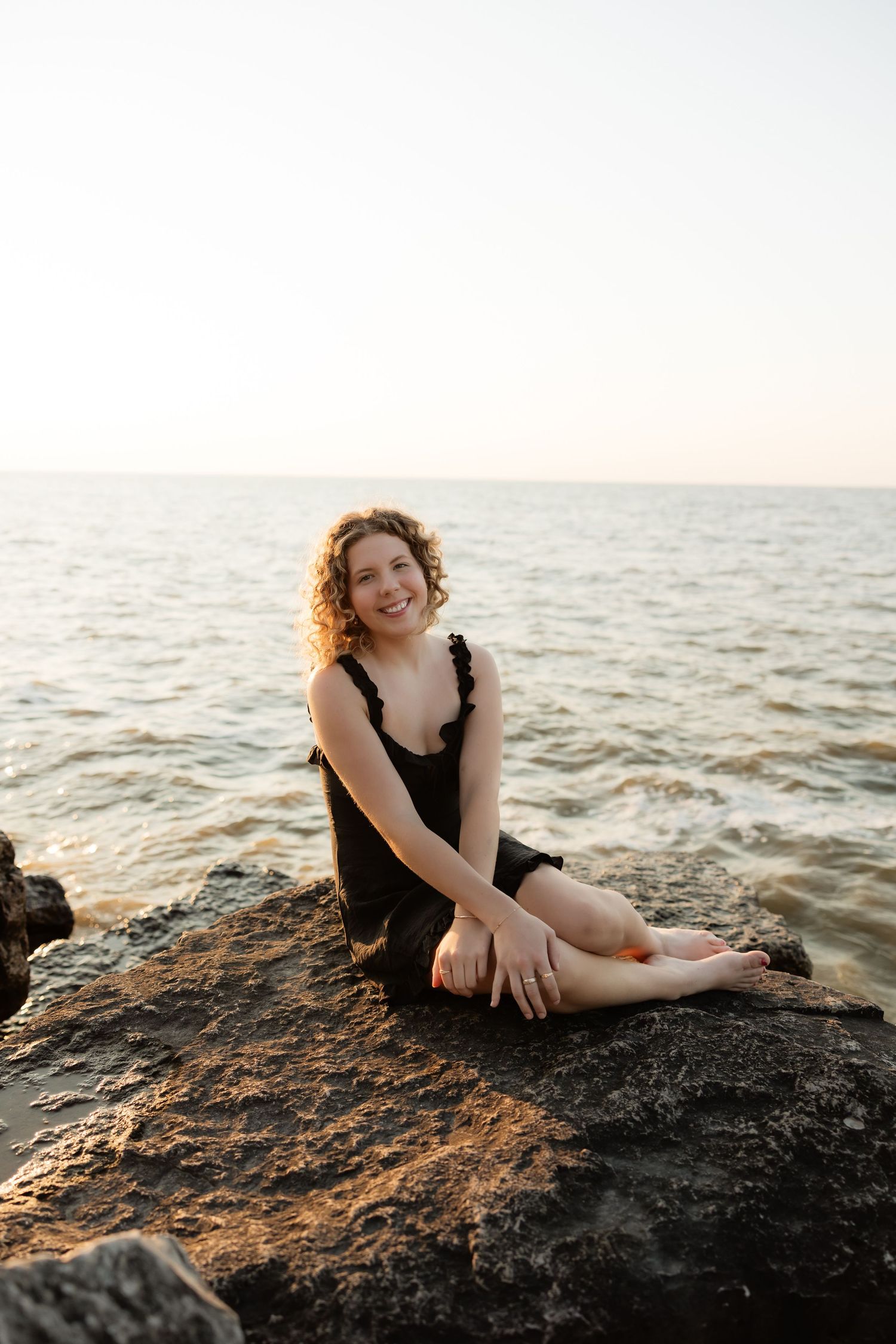Smiling woman with curly hair in black dress sitting barefoot on rocky shore with calm ocean at sunset.