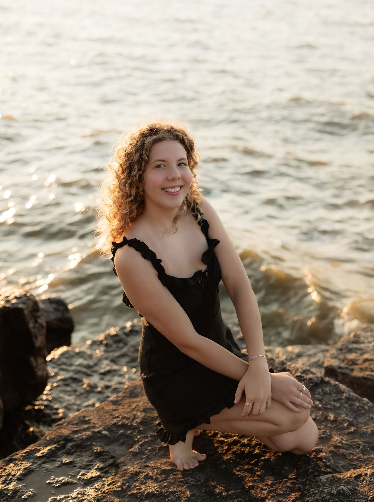 Young woman with curly hair in black dress sitting on rocky shore at golden hour sunset.