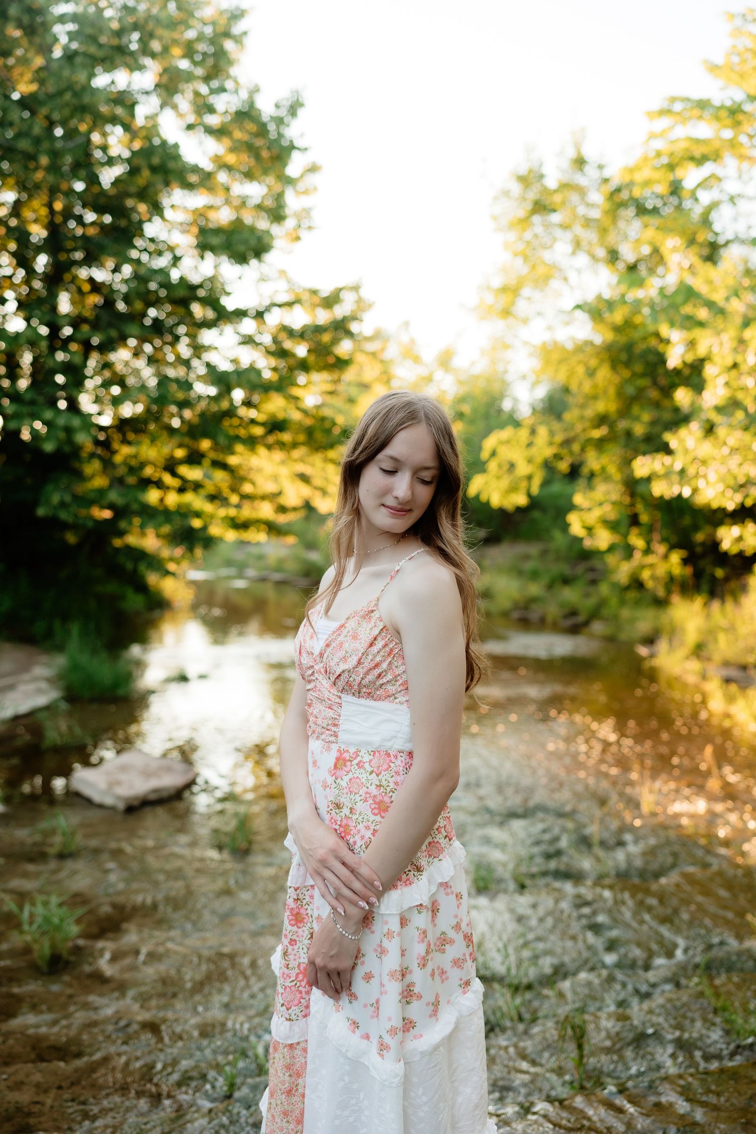 Young woman in floral sundress stands by sunlit creek surrounded by lush green trees.