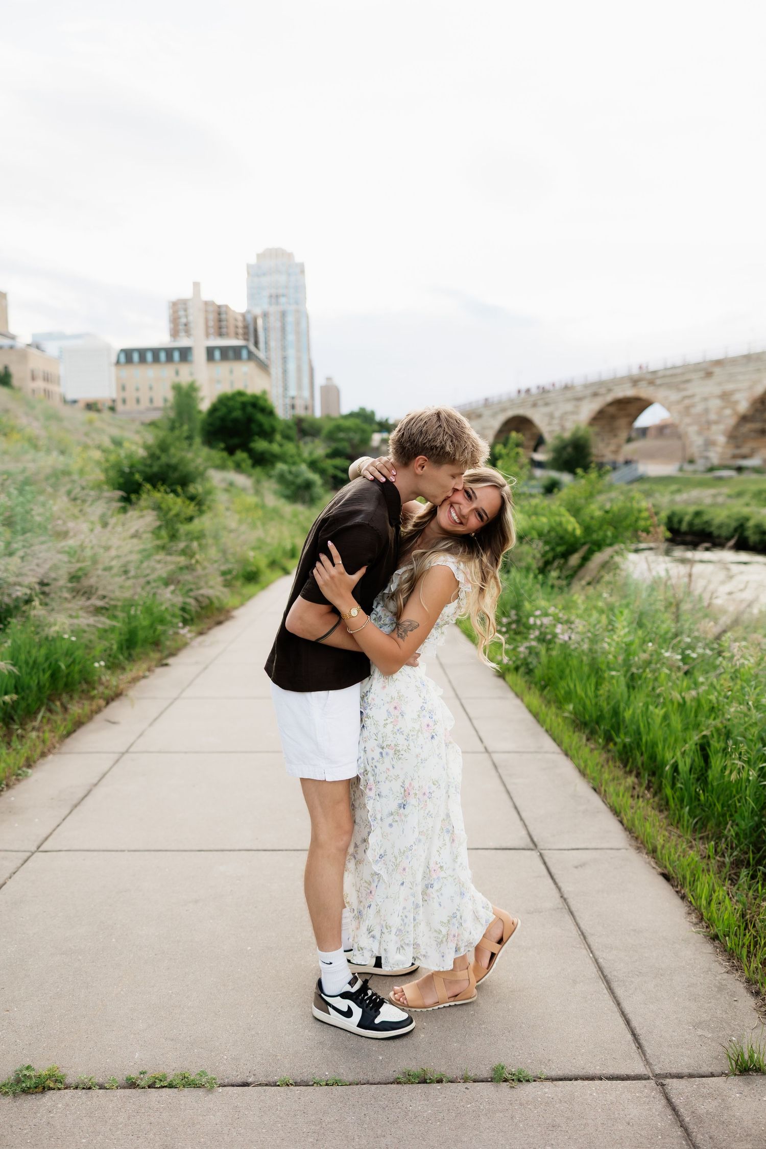 Couple embracing and laughing on a riverside path with a stone arch bridge in the background.