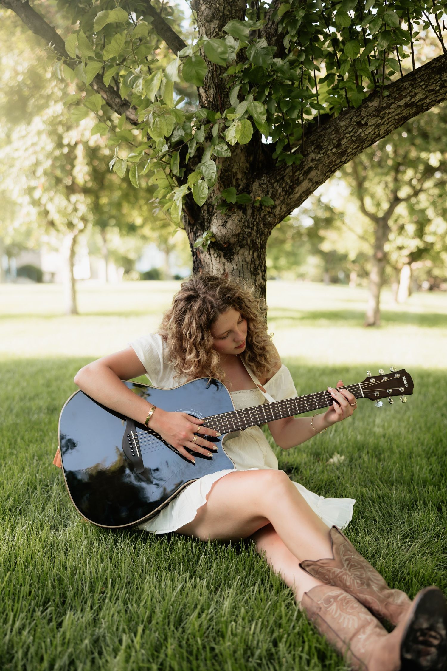 Young woman with curly hair playing acoustic guitar while sitting on green grass under a tree in a sunny park.