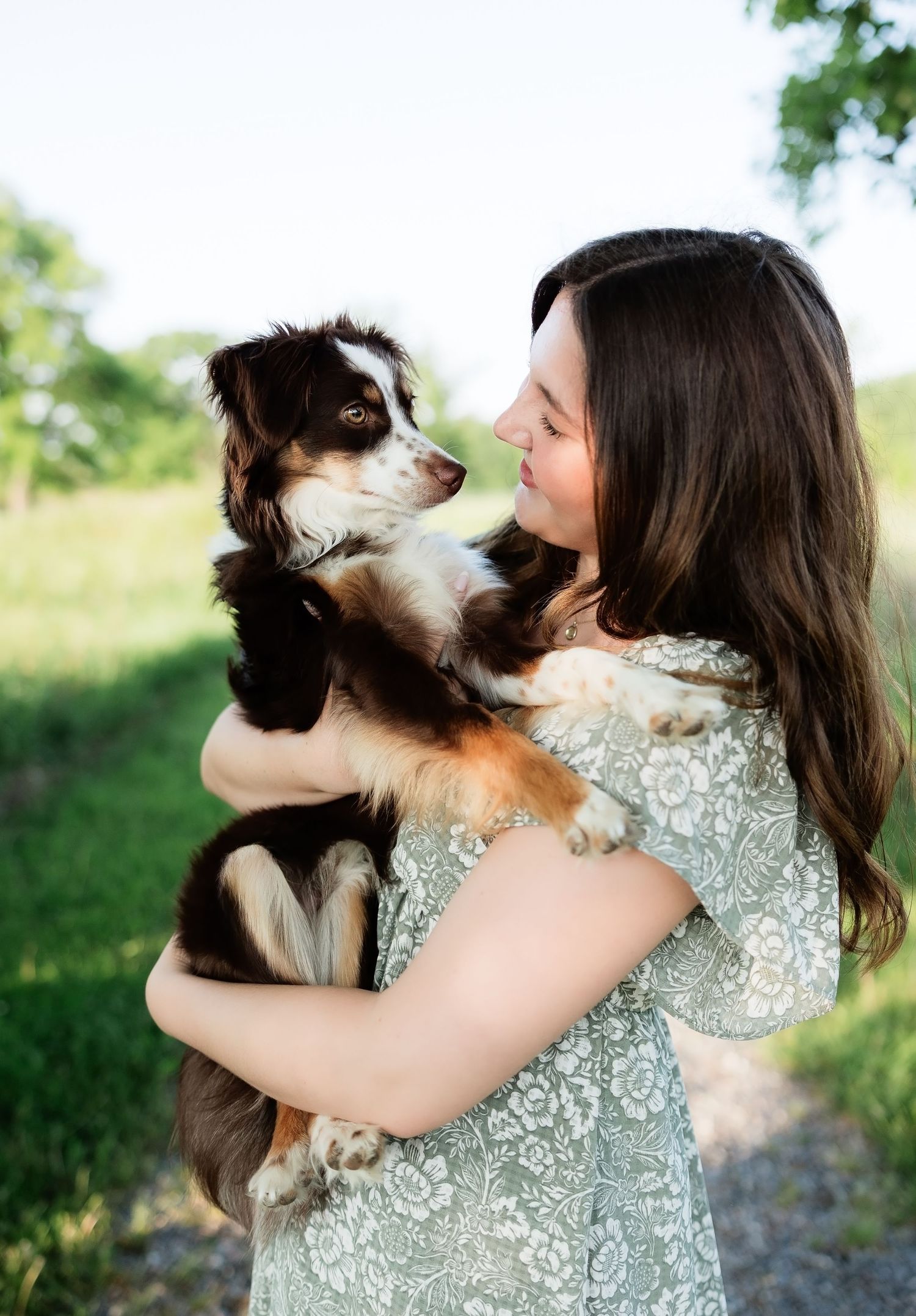 Young woman in floral dress lovingly holding an Australian Shepherd puppy outdoors in natural light.