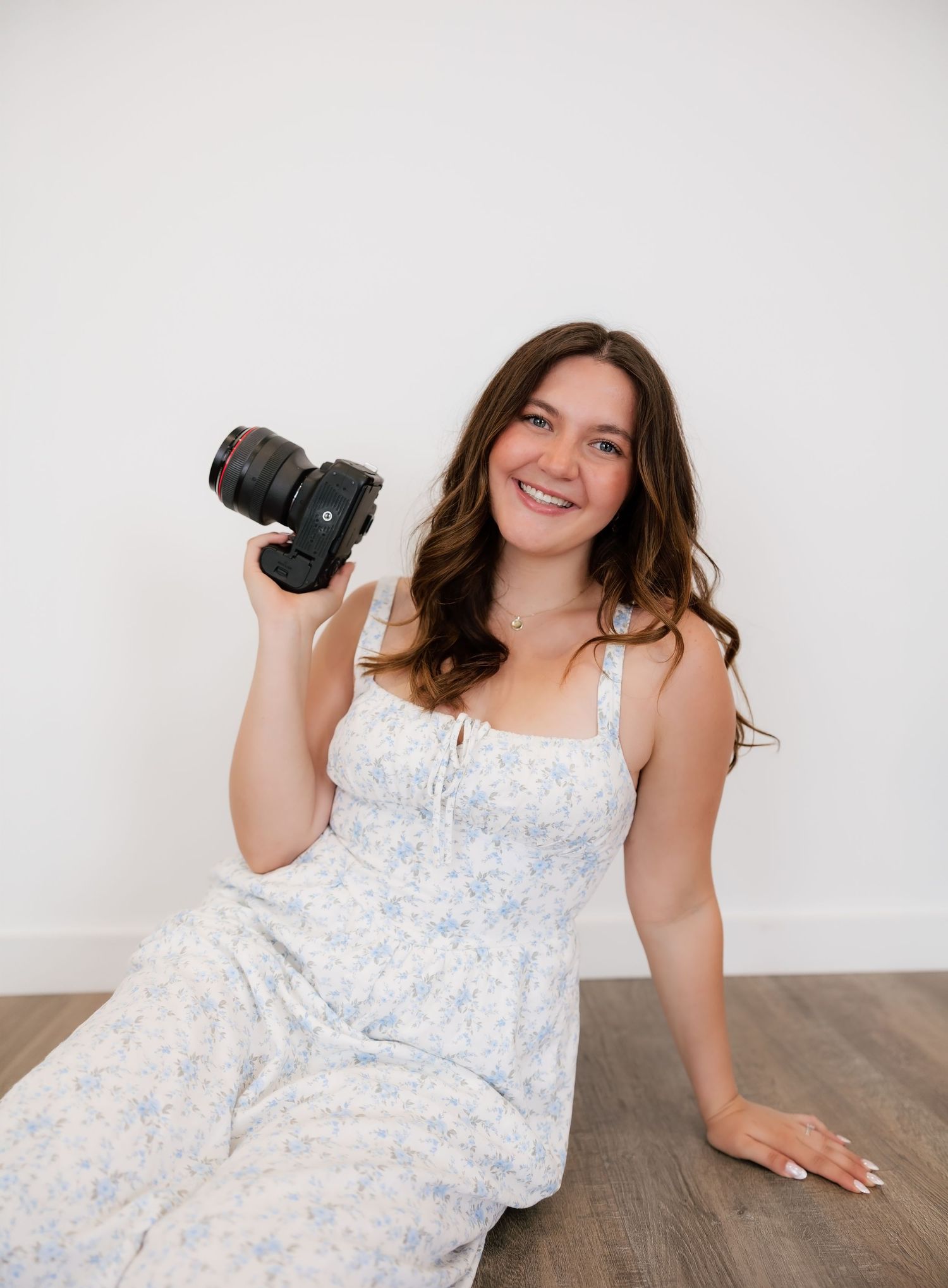 Smiling young woman in floral sundress sitting on hardwood floor holding a DSLR camera against white wall.