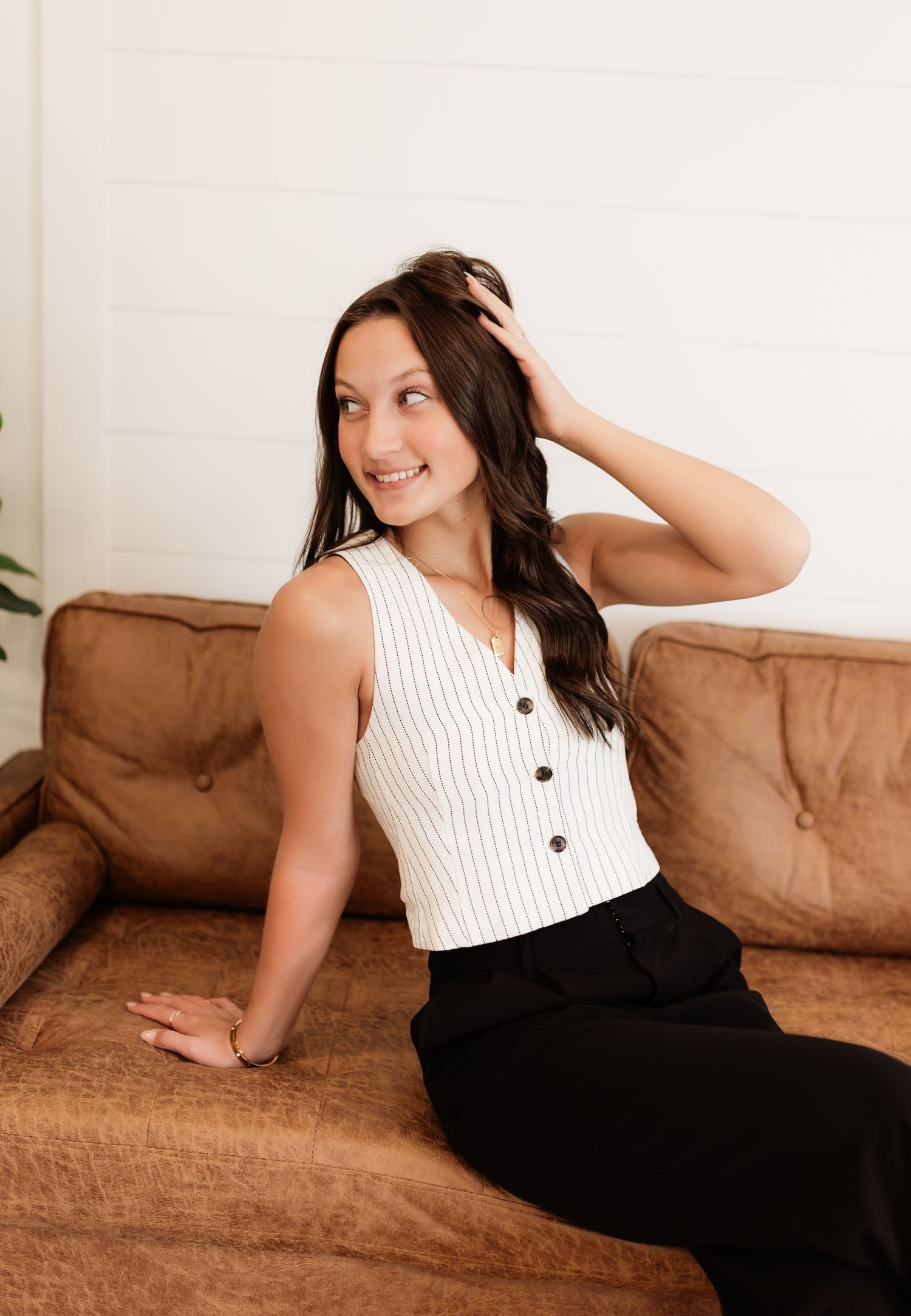 Young woman in white striped vest and black pants smiling on brown leather couch.