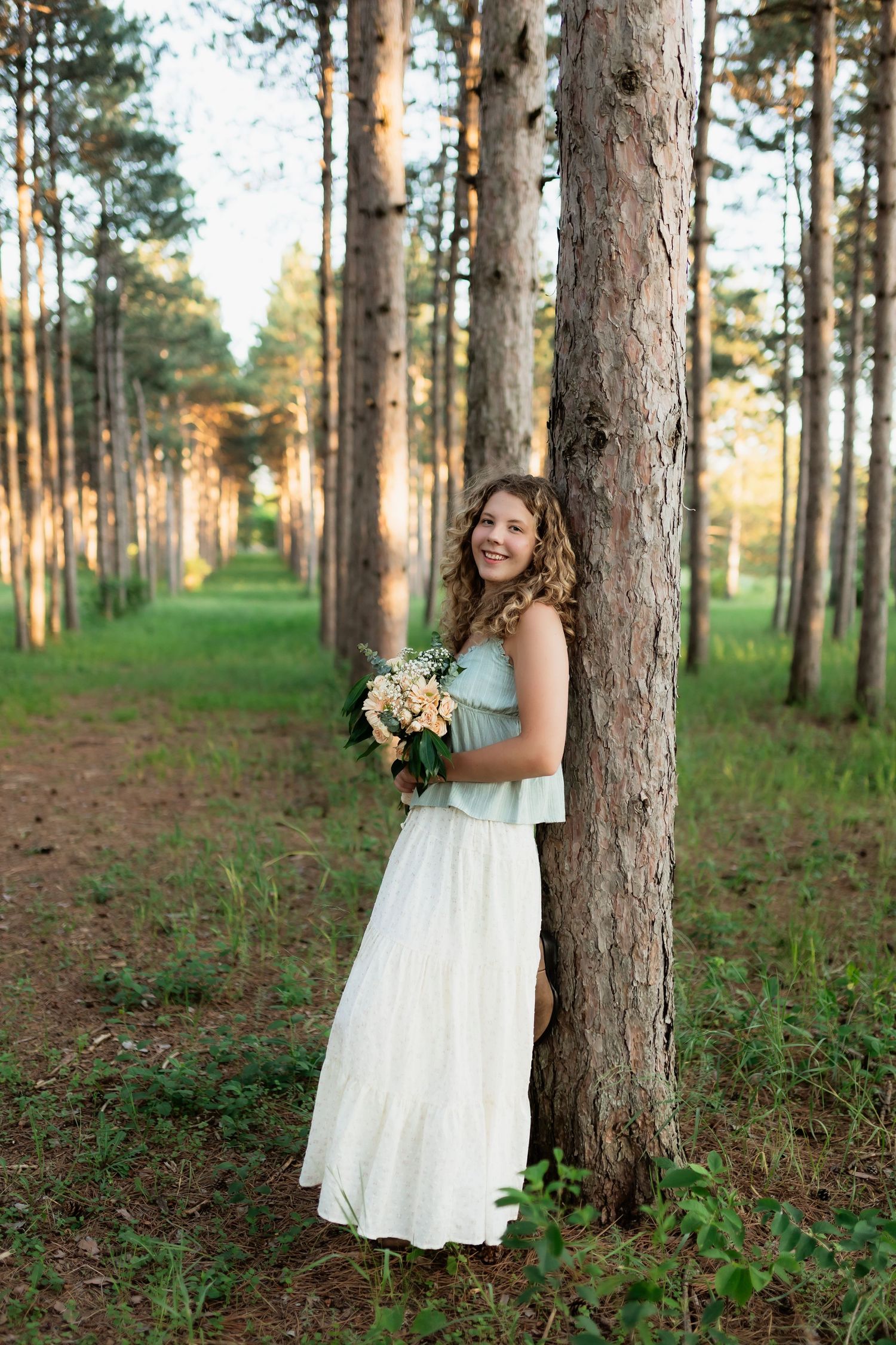 Smiling young woman with curly hair holding a floral bouquet while leaning against a pine tree.