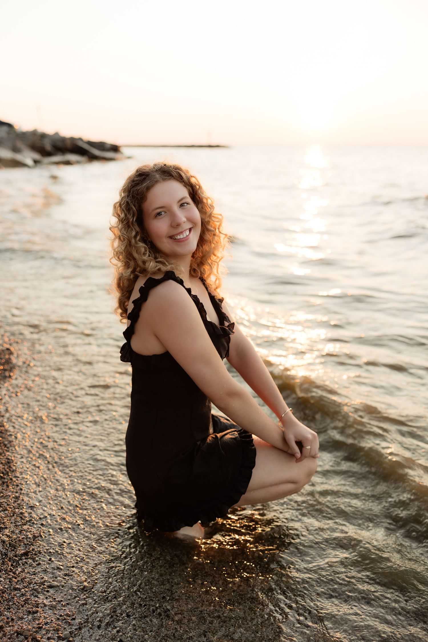 Young woman with curly hair in black dress smiling at beach sunset, crouching in shallow water.