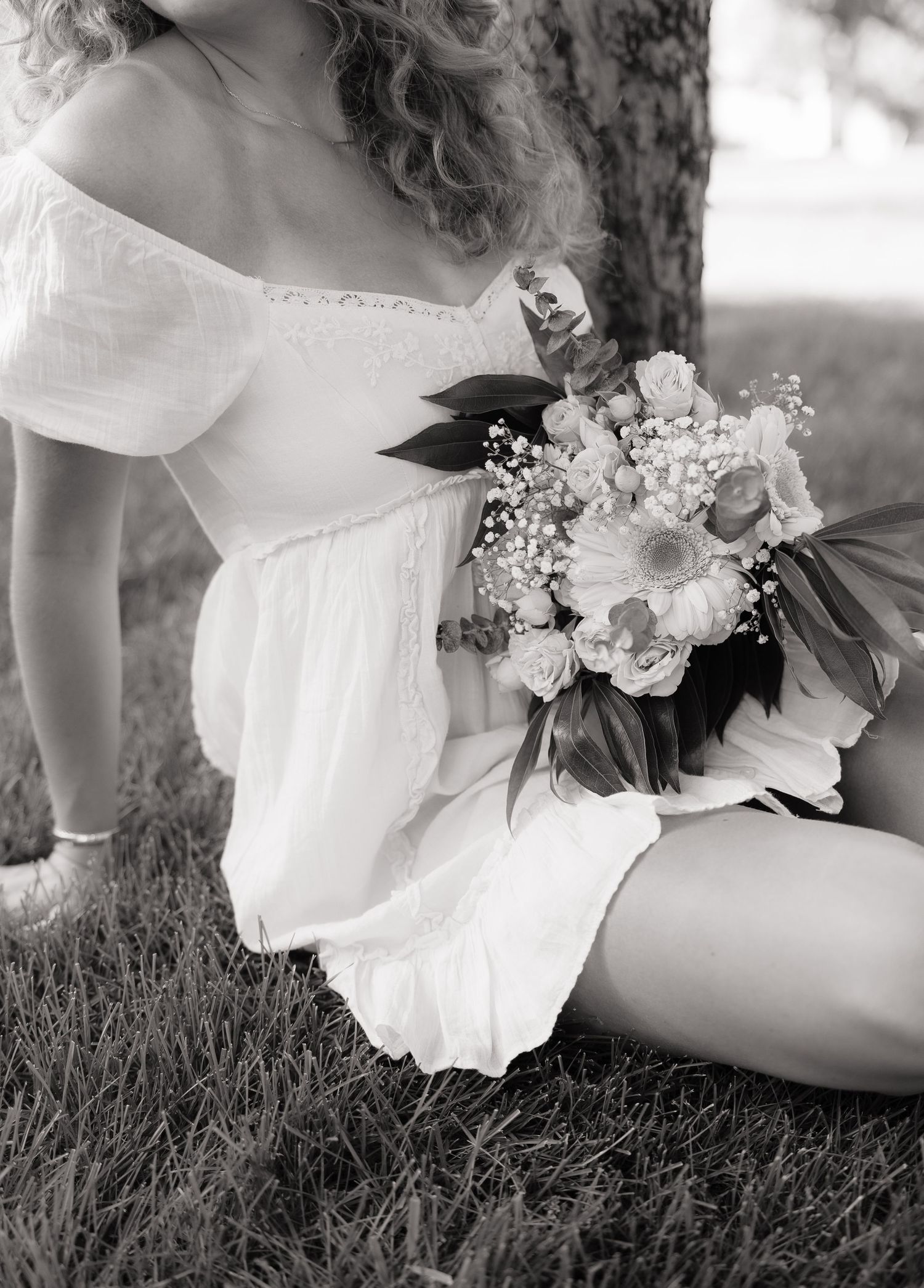 Black and white photo of a woman in a white off-shoulder dress sitting by a tree holding a floral bouquet.